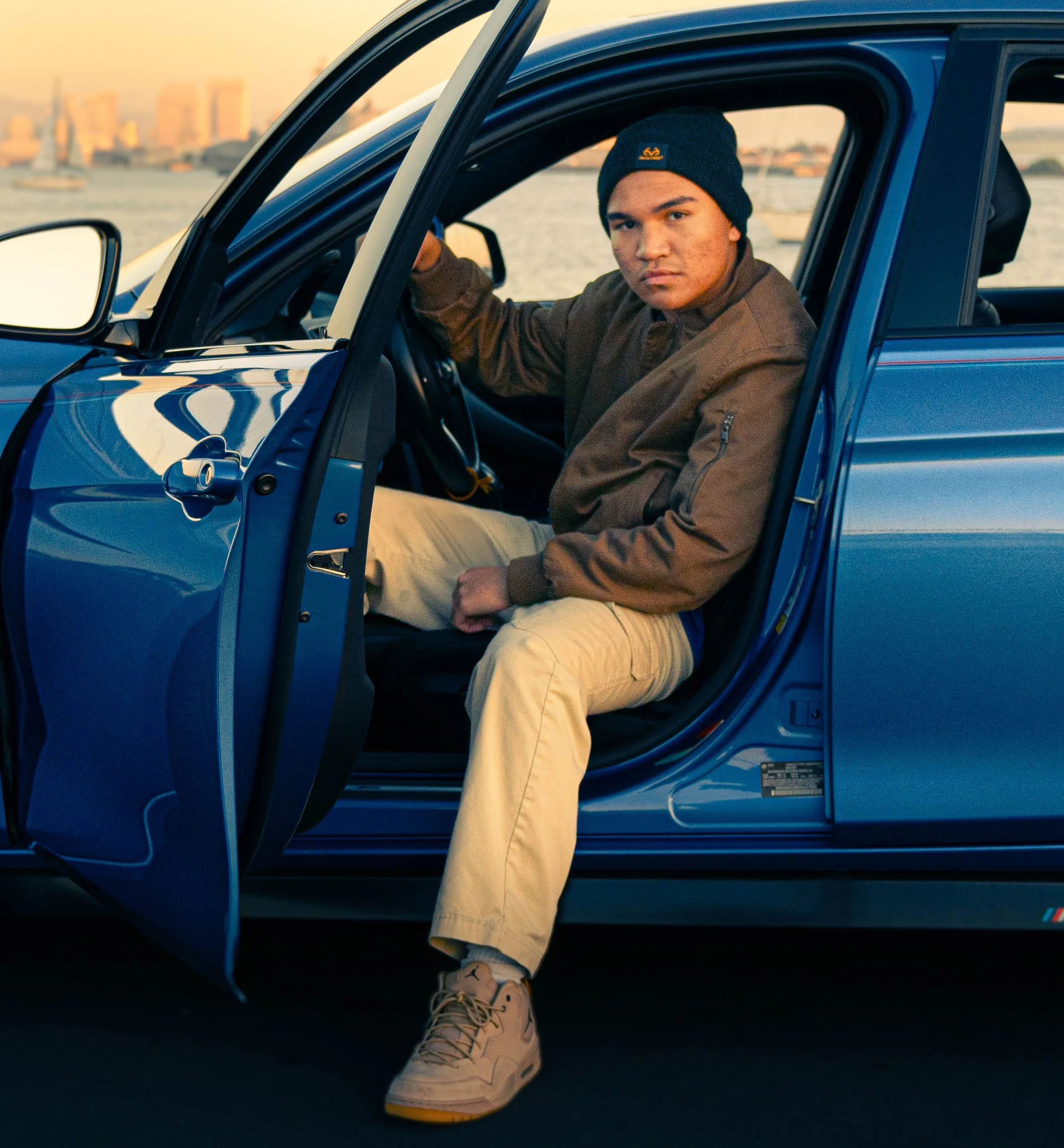 A young man with tan skin, wearing a dark beanie and brown jacket, is sitting in the driver's seat of a blue car with the door open. His left arm is resting on the car door, and he is wearing beige pants and white sneakers. The image is taken at suns