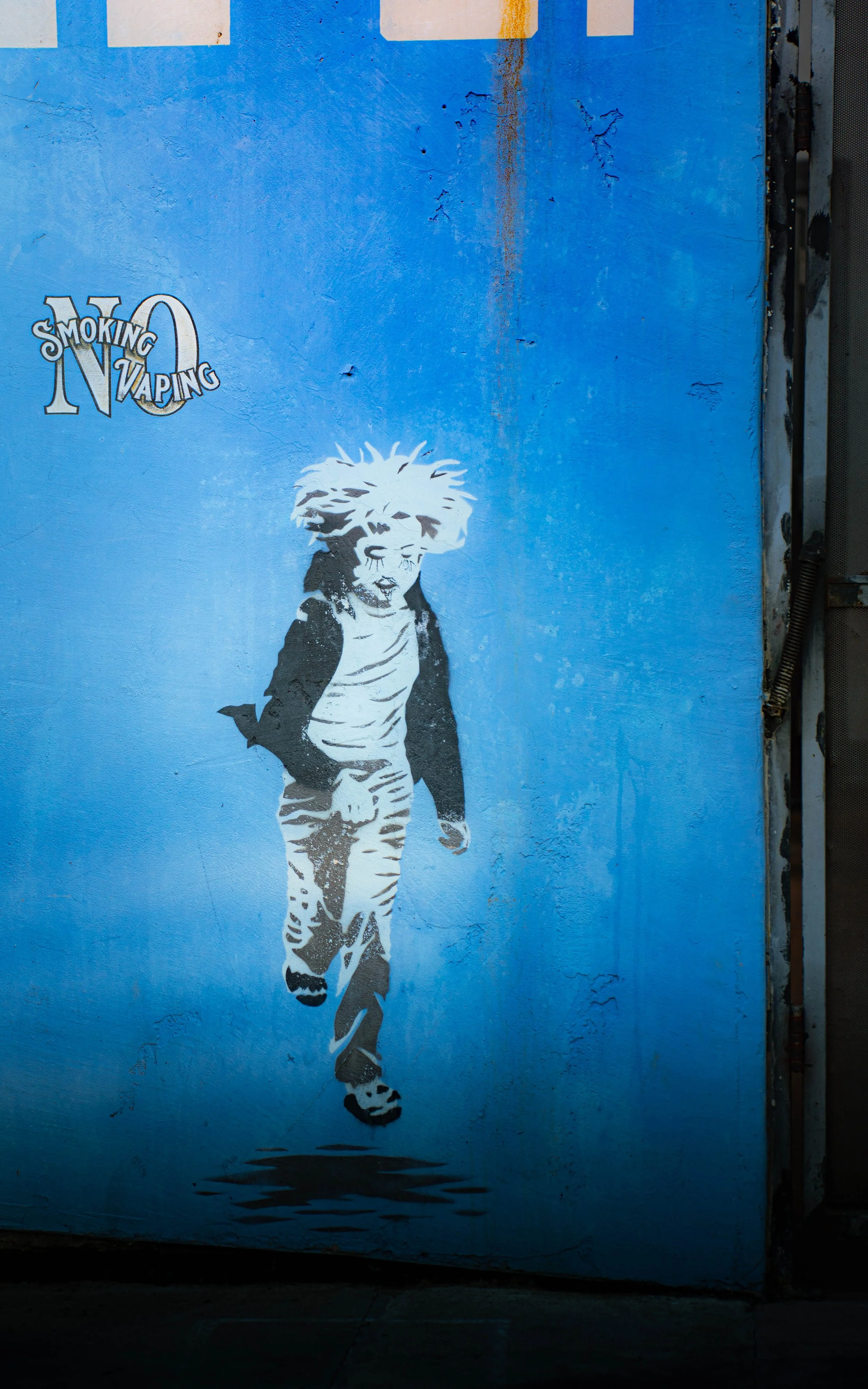 Street art graffiti of a child with spiky hair, wearing a jacket, t-shirt, and striped pants, walking on a blue wall with a no-smoking and no-vaping sign.