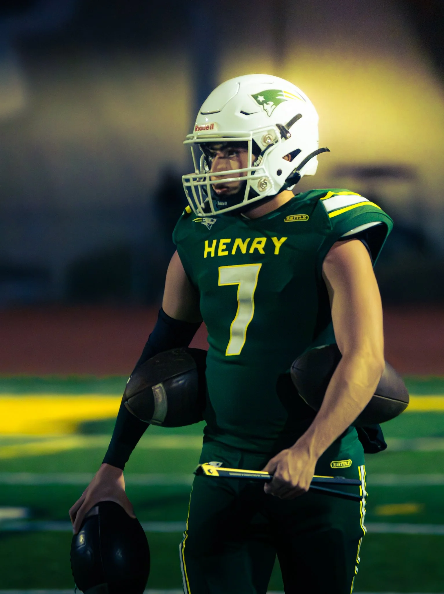 A football player wearing a white helmet with a New England Patriots logo and a green jersey with yellow lettering and stripes, standing on a football field at night.