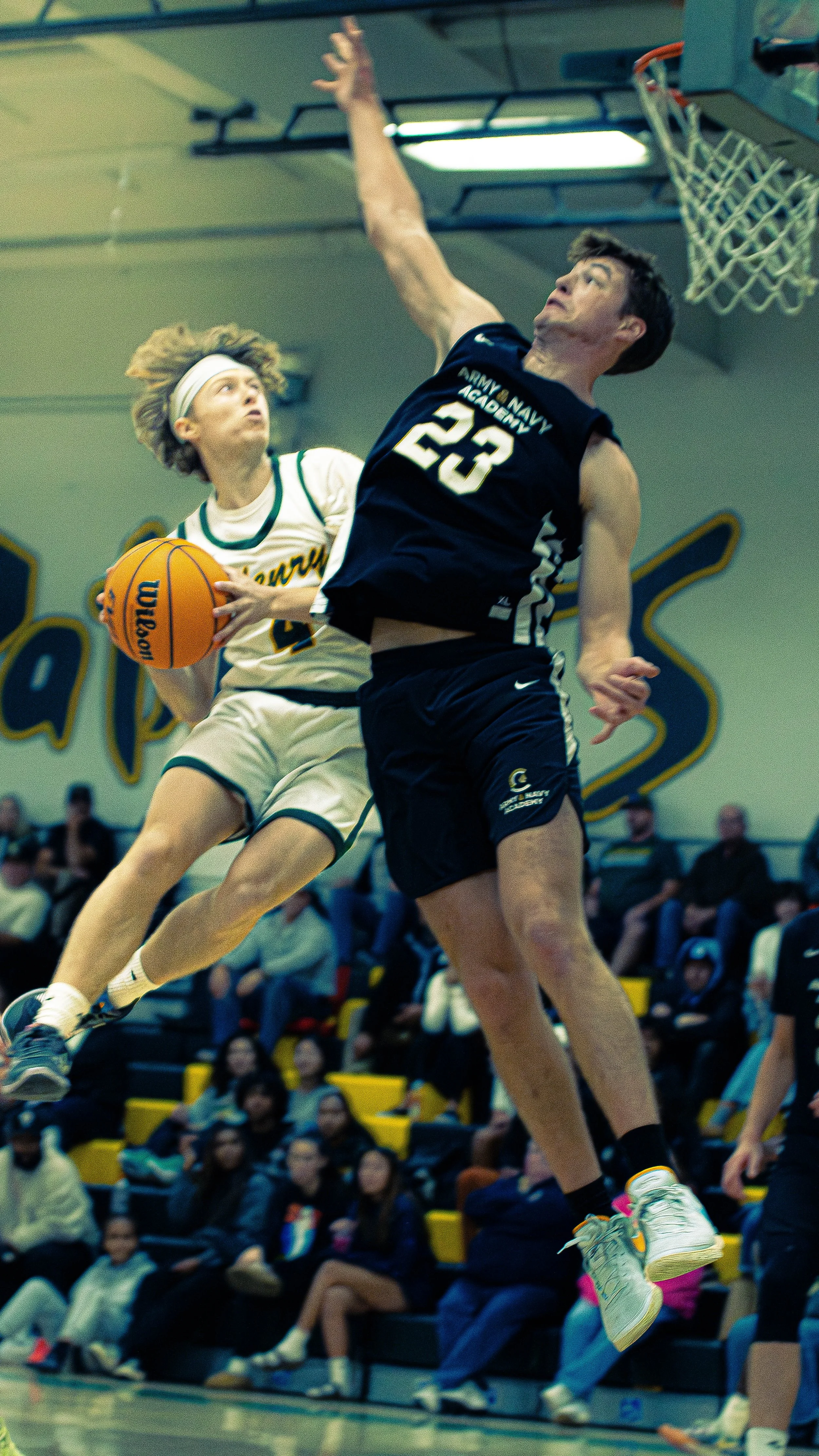 Two young male basketball players jumping for a rebound during a game in a gymnasium. One player is wearing a white jersey with green accents and the other is in a dark blue jersey with white accents. Spectators are seated in the background.