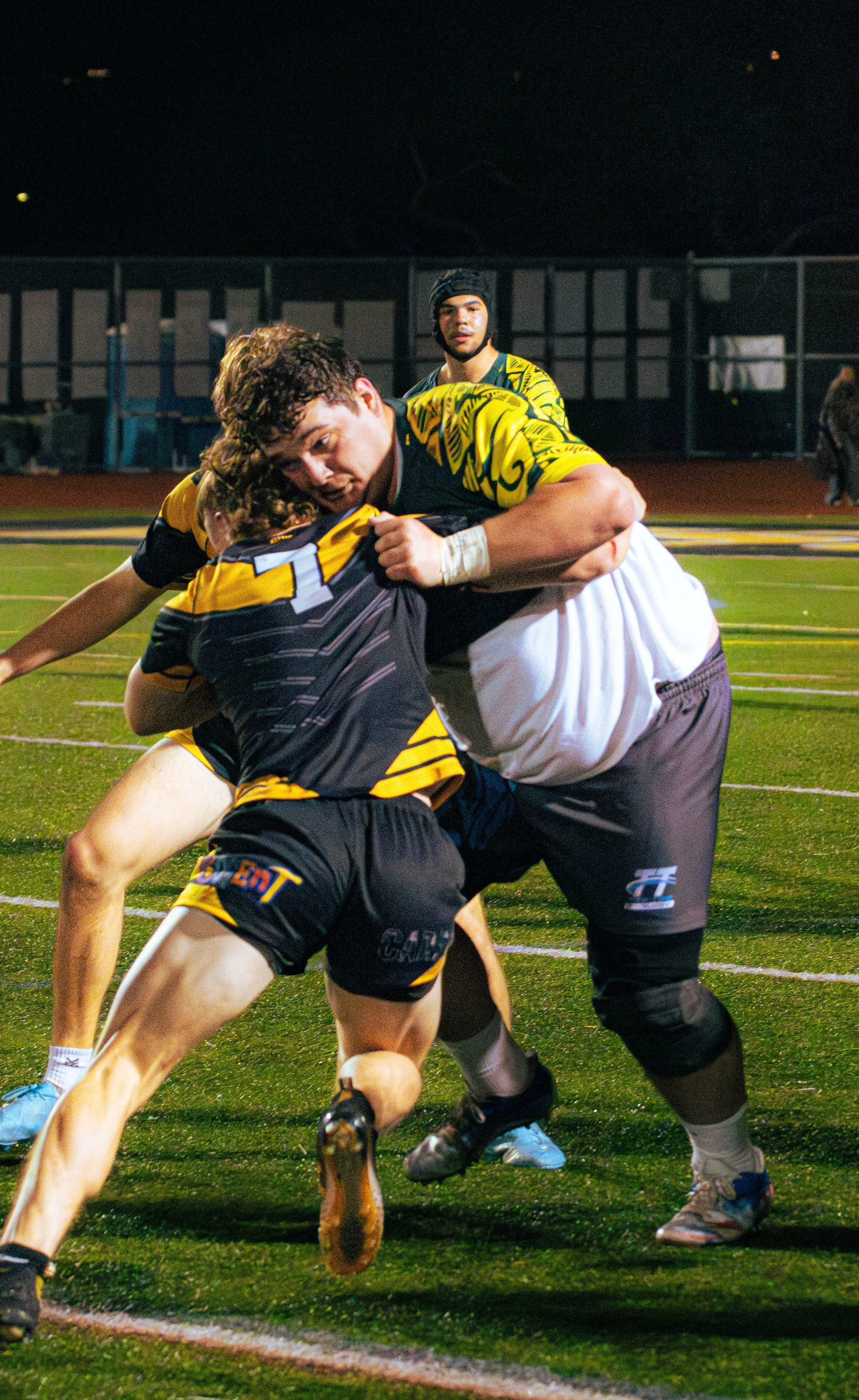 Rugby players engaged in a tackle during a match on a field at night, with two players in black and yellow uniforms and one in a green and yellow uniform.