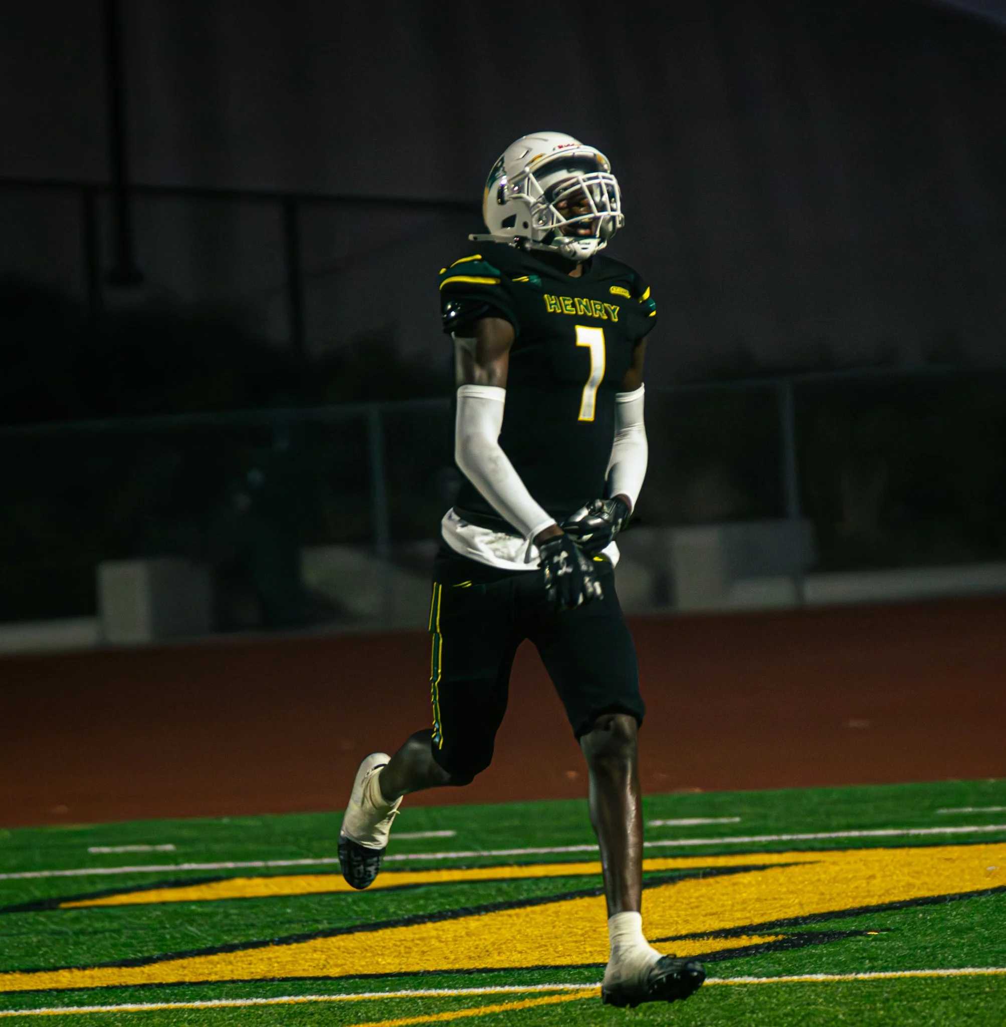 A football player from Henry High School wearing a black jersey with yellow and green accents, white gloves, and a white helmet with a faceguard, running on a green and yellow sports field during a game.