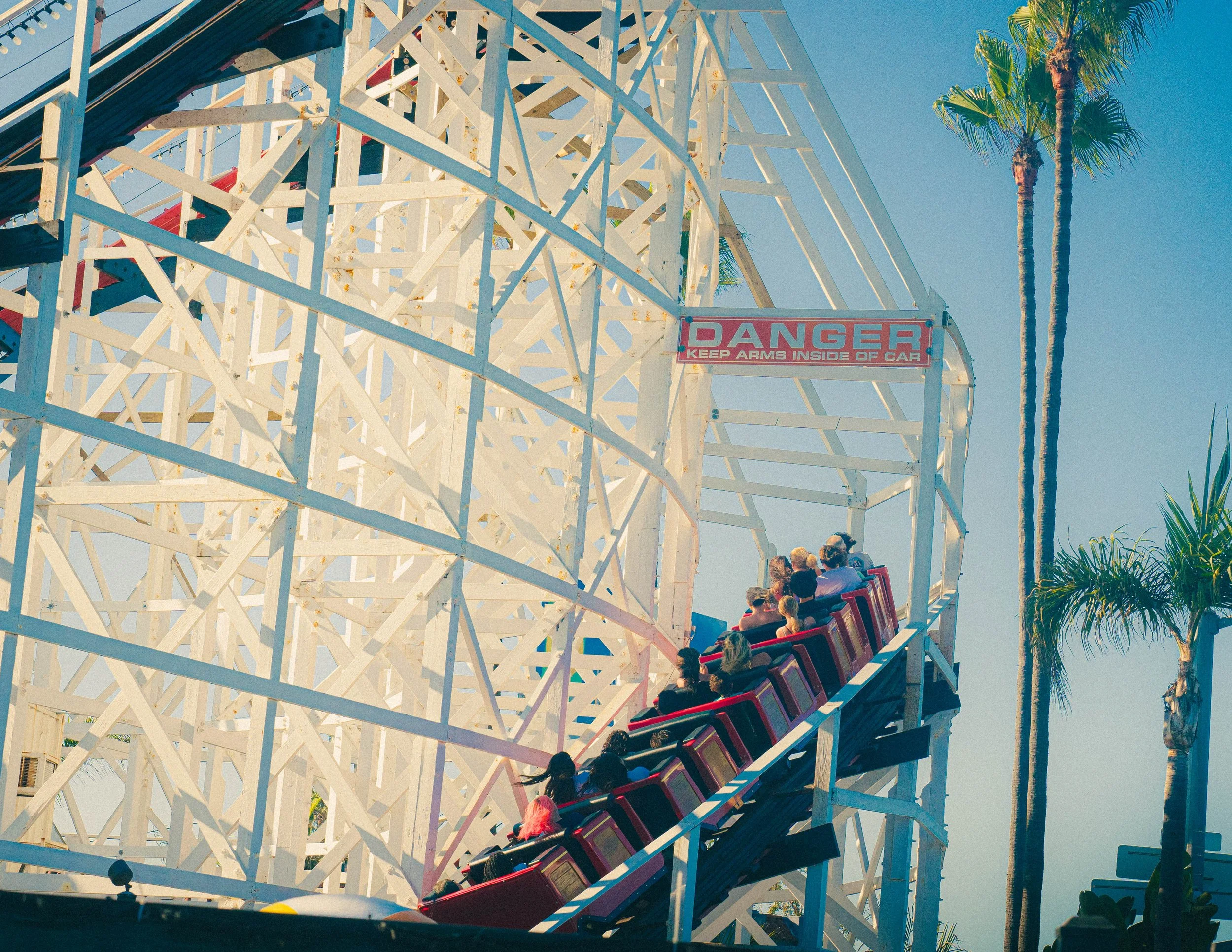 People riding a colorful wooden roller coaster at an amusement park with palm trees and a clear blue sky in the background.