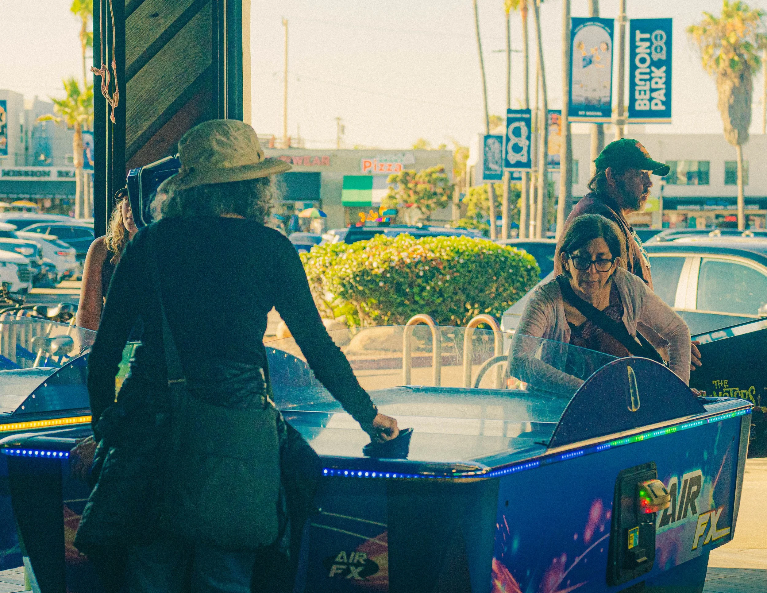 People on an arcade ride shaped like a vintage car with colorful LED lights, with a background of signs and buildings in a shopping center parking lot.