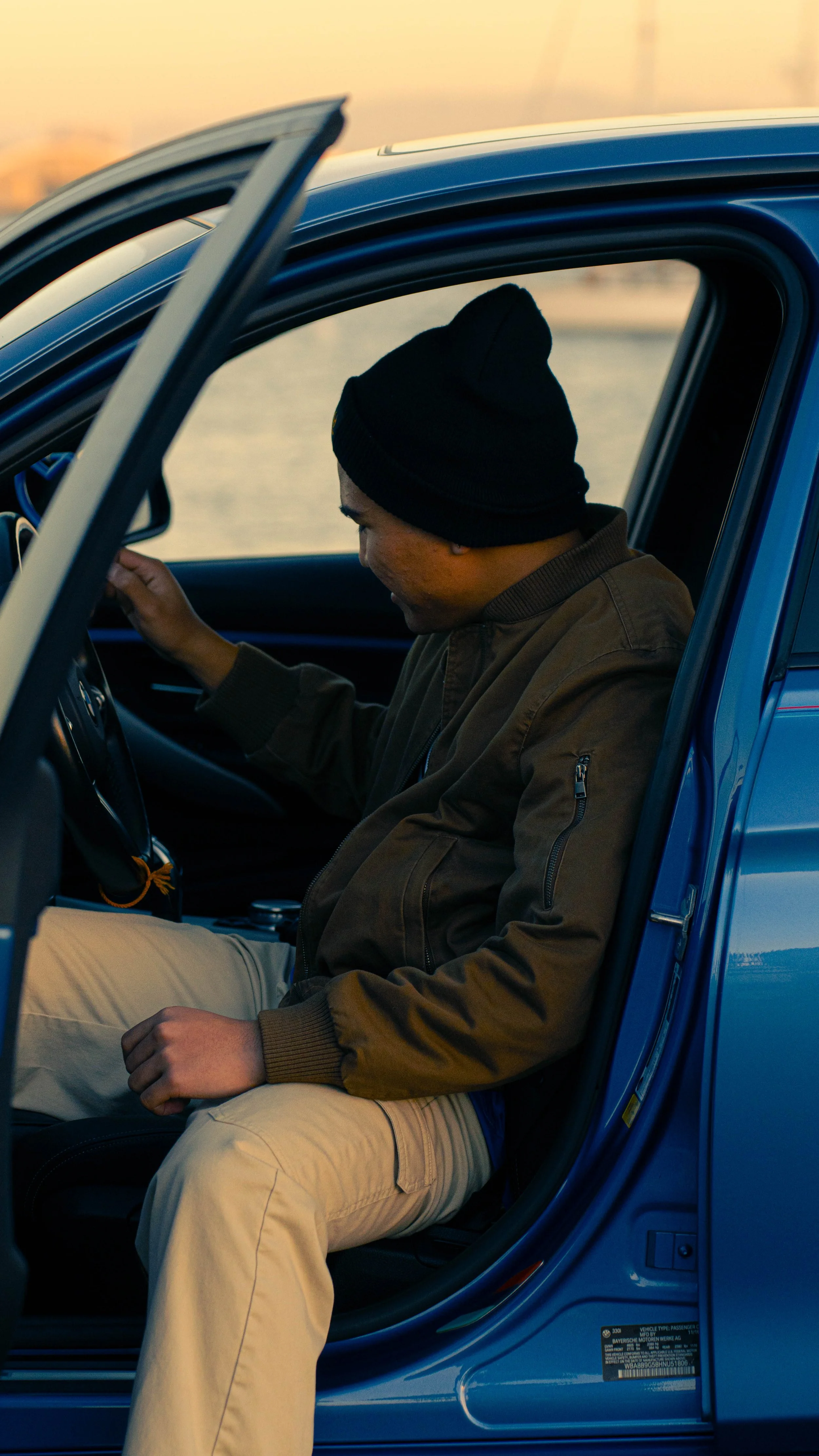 Young man wearing a black beanie and brown jacket sitting in a blue car, looking down at his hands, during sunset.