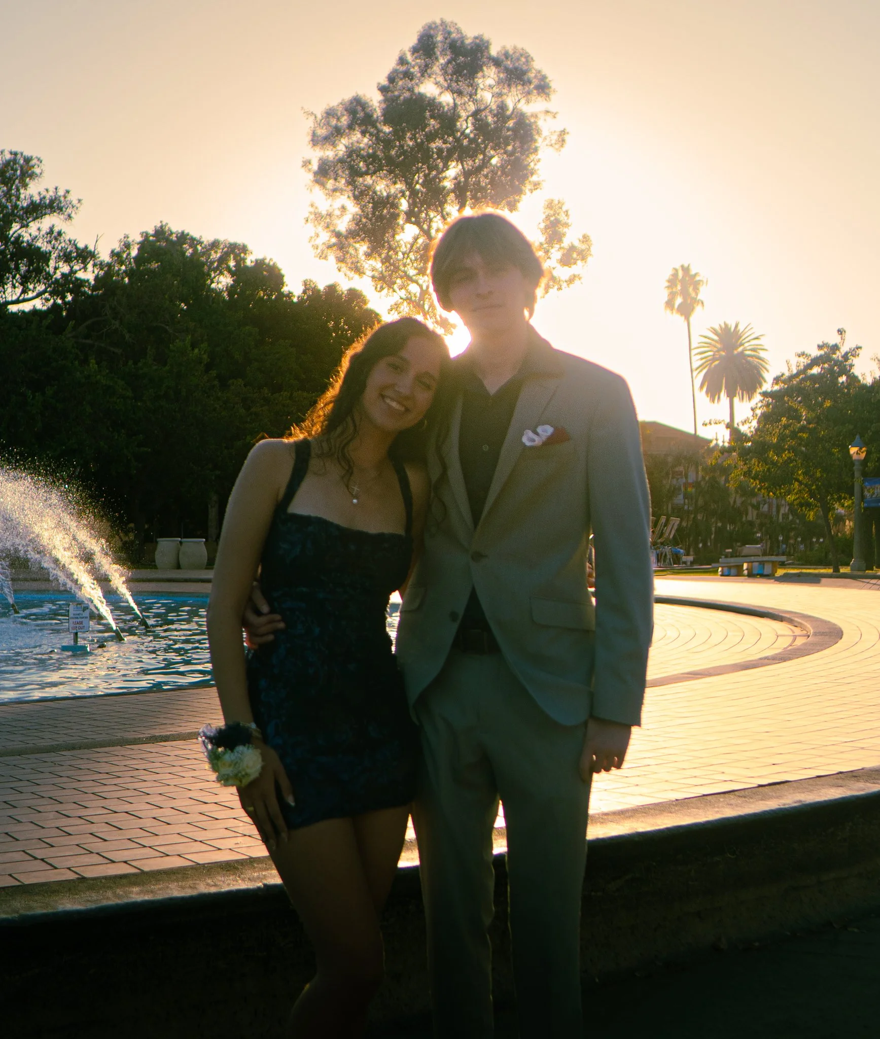 A young woman and a young man posing together outdoors during sunset, with trees in the background. The woman is holding a bouquet of flowers and smiling, while the man is wearing a light gray suit with a lapel pin and a white pocket square.