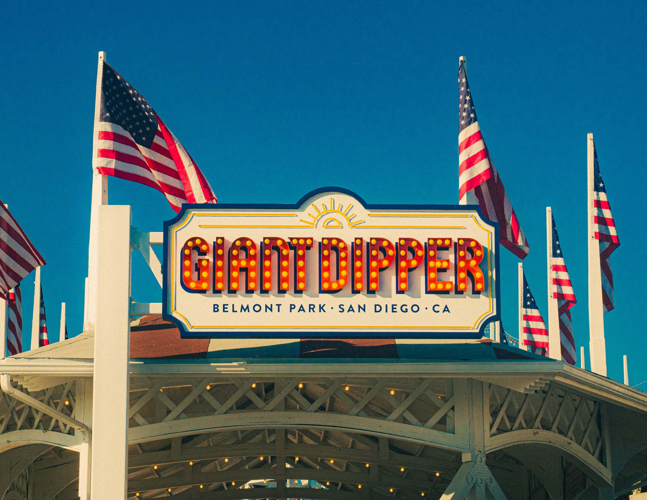 Sign that reads 'GARDEN DIPPER' with the location 'BELMONT PARK - SAN DIEGO, CA' beneath it, flanked by American flags on poles against a clear blue sky.