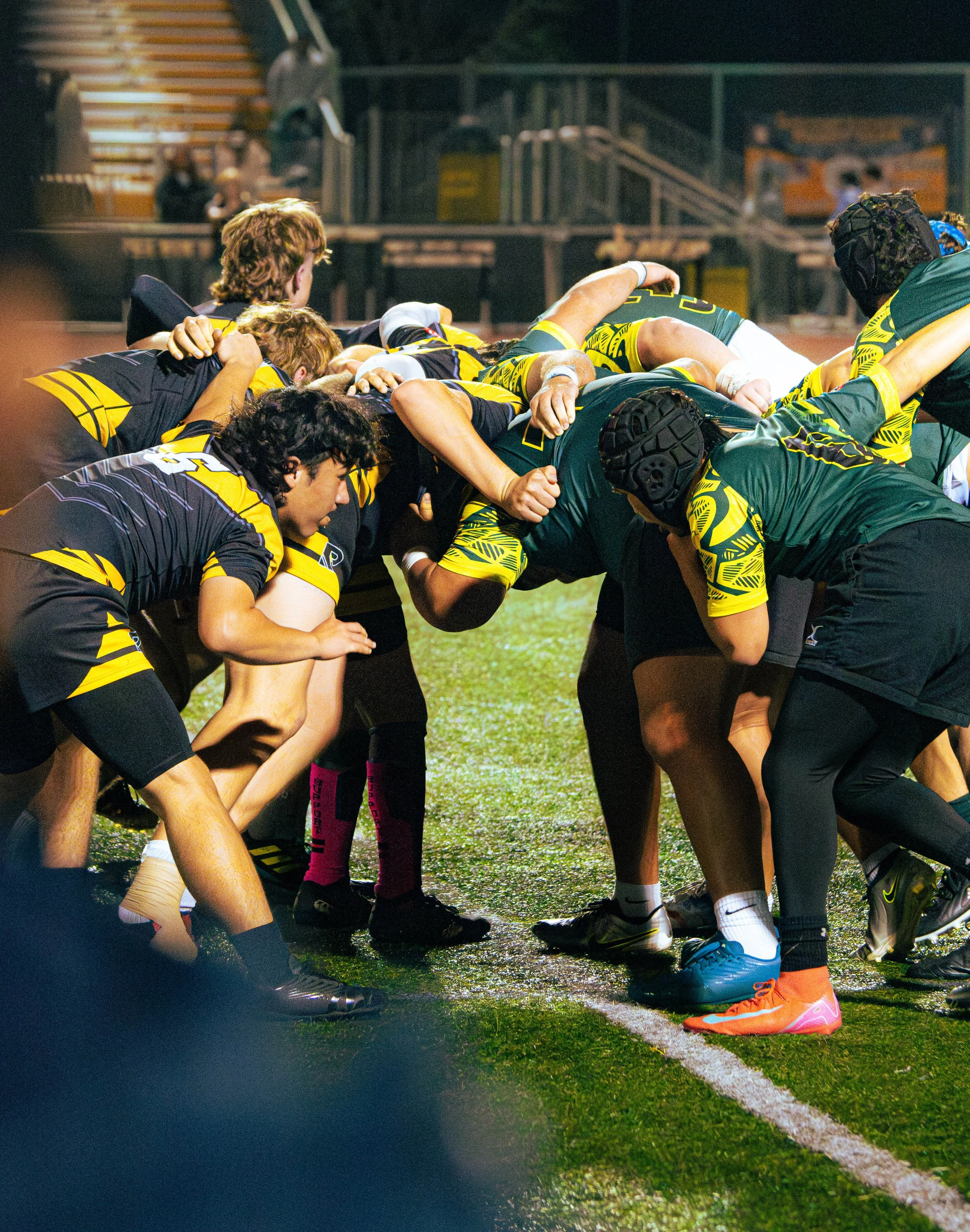 Rugby players forming a scrum on the field at night, wearing black, green, and yellow uniforms.