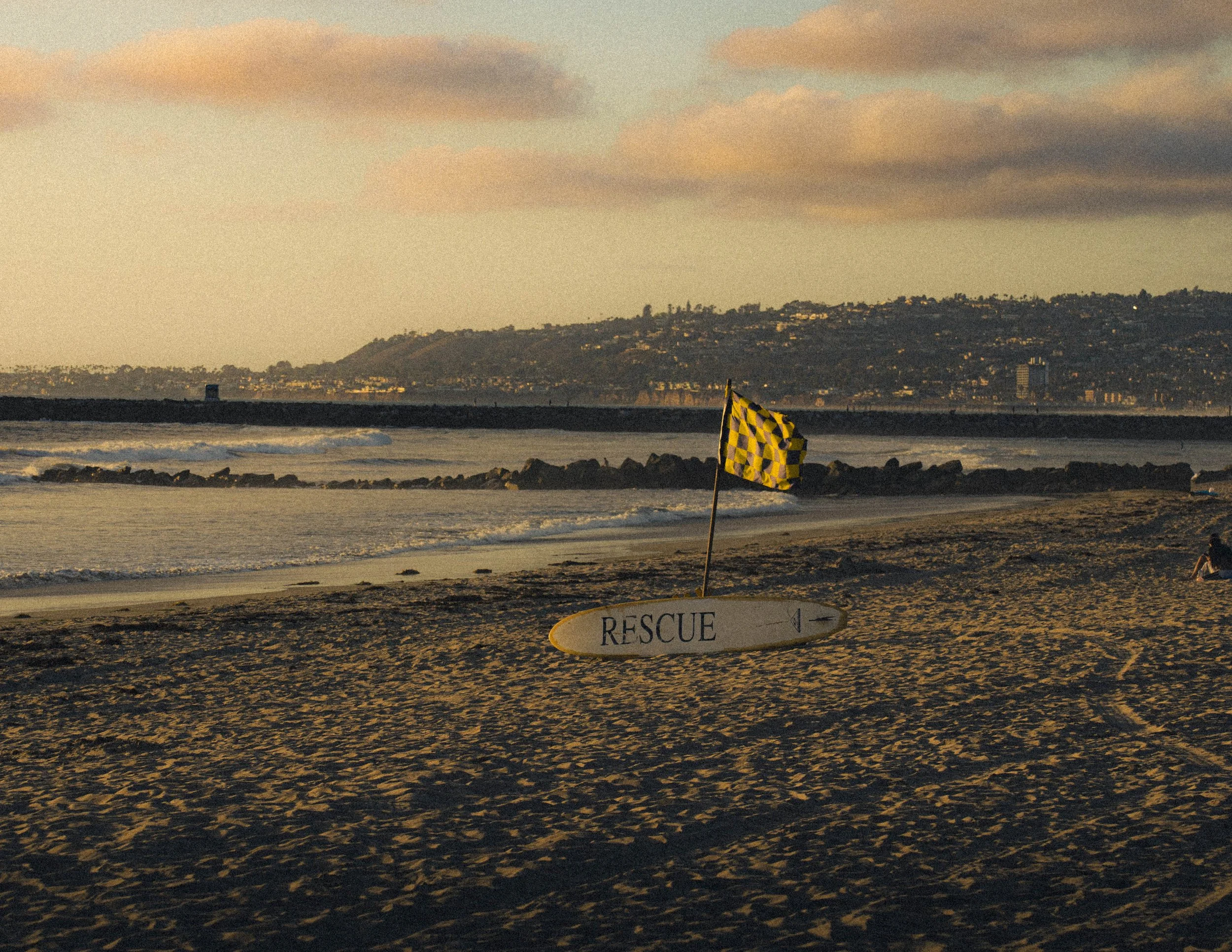 A beach at sunset with a yellow and black checkered caution flag in the sand, and an oval rescue sign lying on the ground. In the background, there is a body of water with waves and a distant hillside with houses and a few buildings, under a partly c