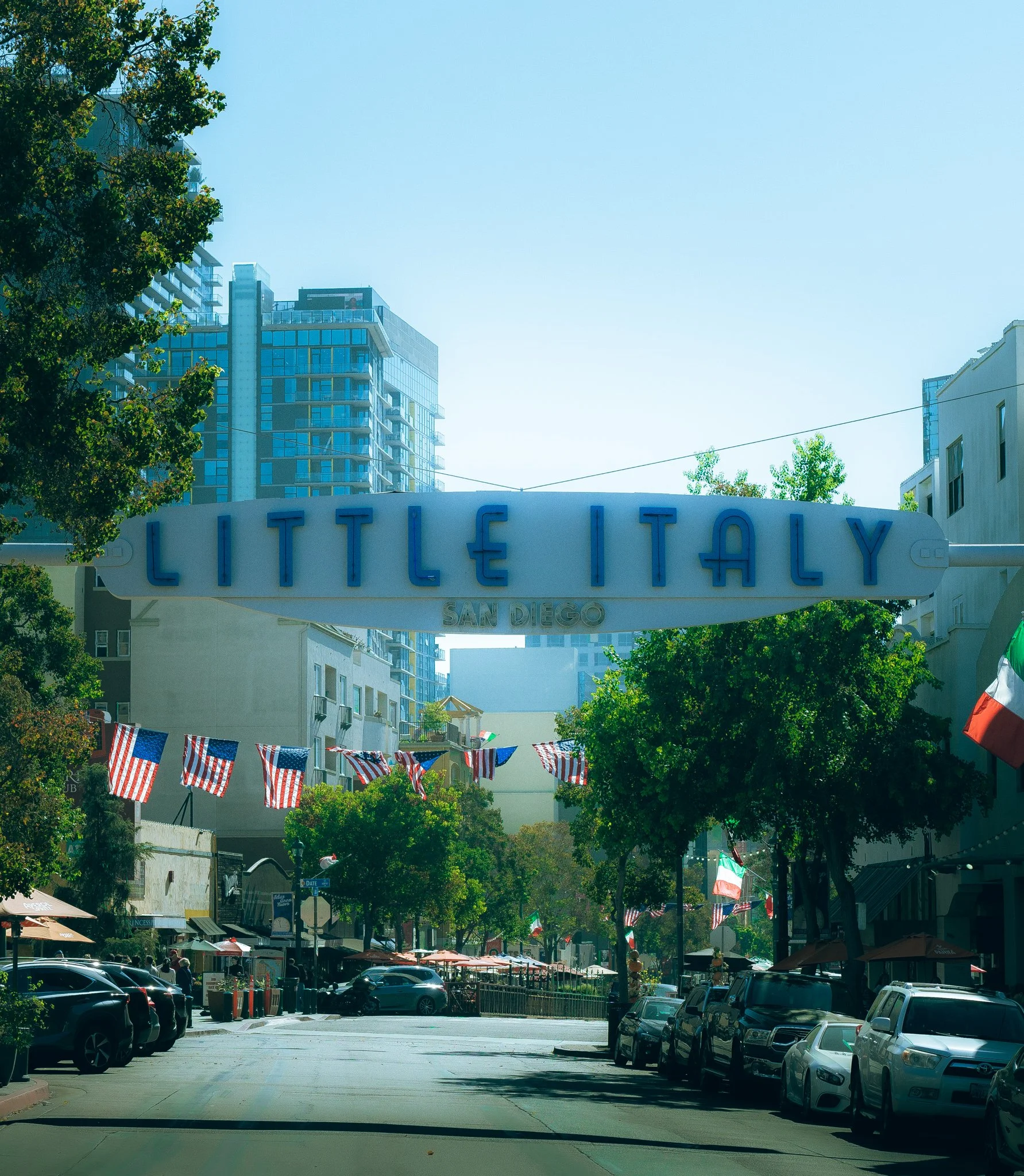 A busy street in Little Italy, San Diego, decorated with American flags, with parked cars and outdoor cafes, and a large sign overhead reading 'Little Italy San Diego'.