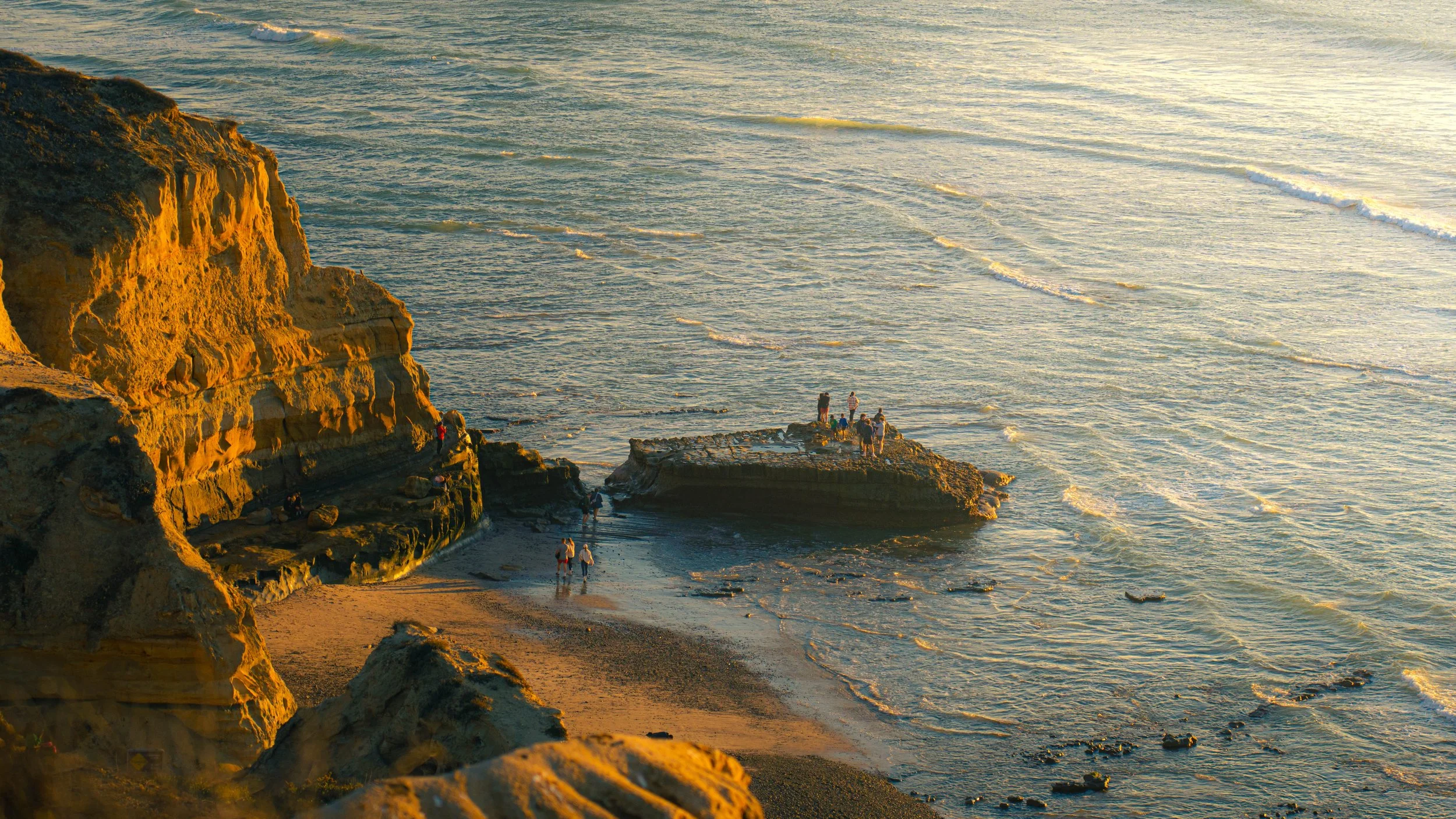 Golden sunset over rocky coast with people gathering on a small island in the ocean.