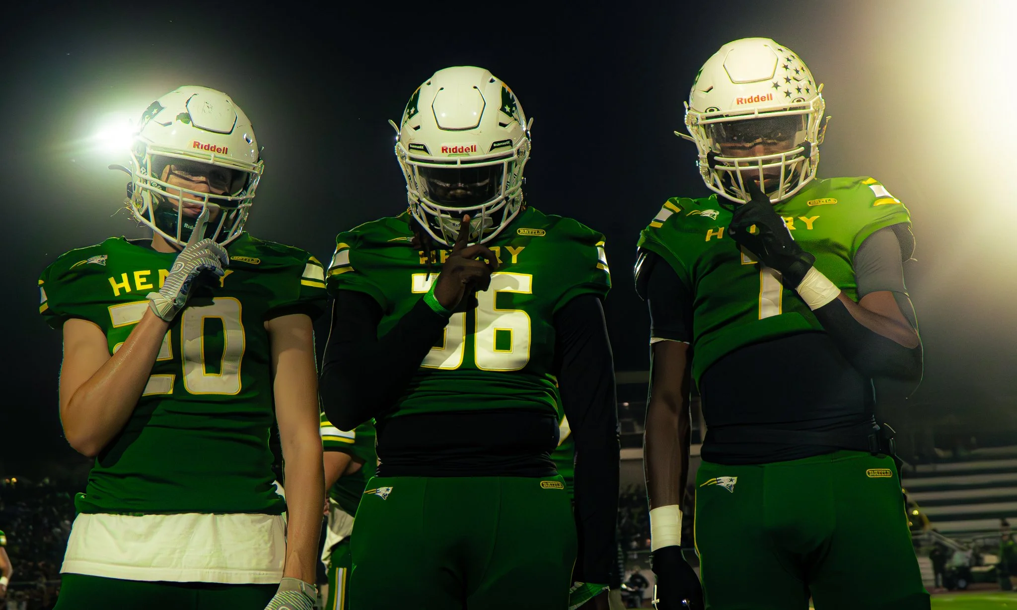 Three American football players wearing green jerseys and white helmets, posing on the field at night, with their index fingers raised to their lips in a silencing gesture.
