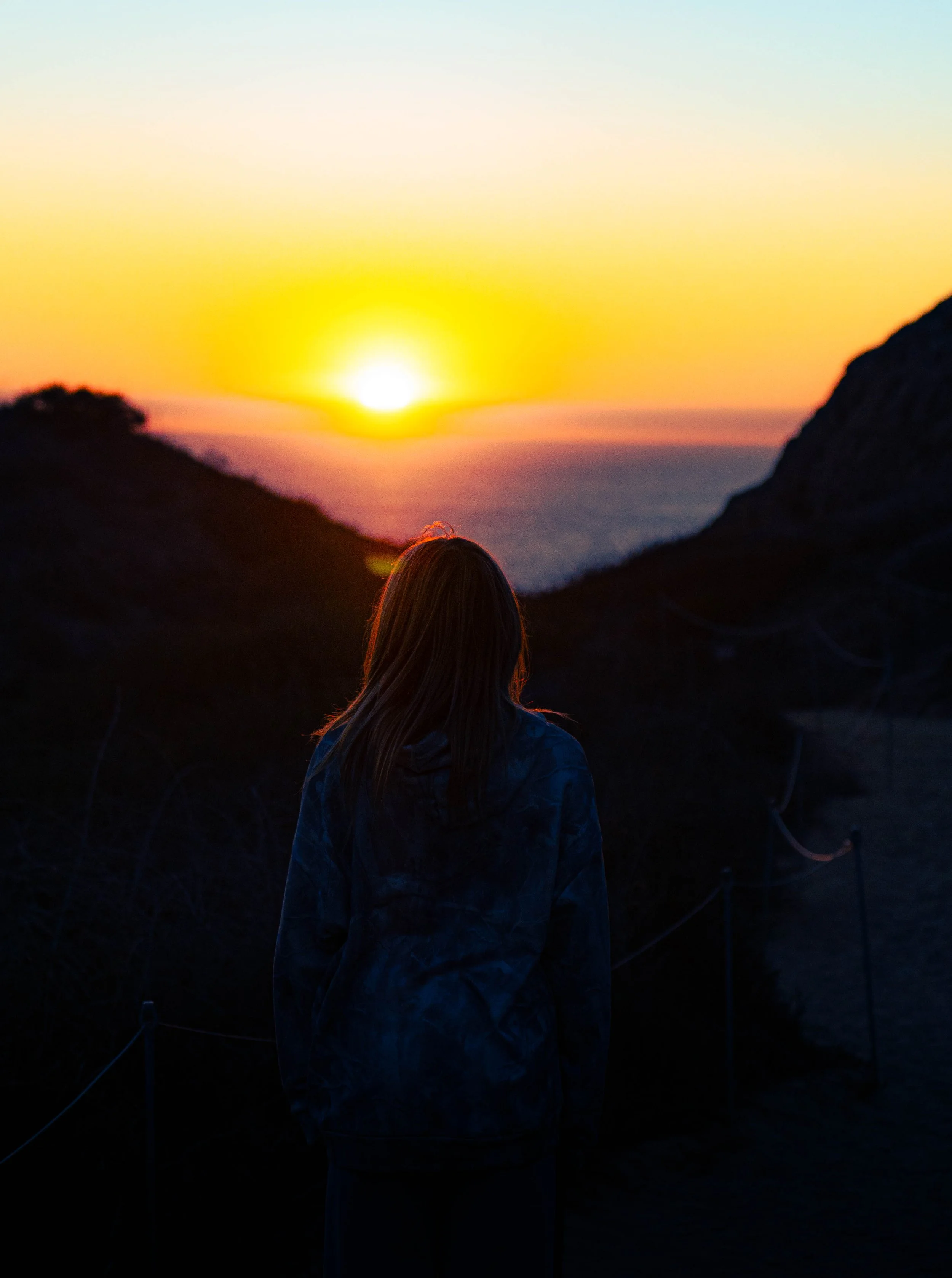 A person with long hair wearing a dark jacket standing outdoors at sunset, overlooking a landscape with mountains and the ocean.