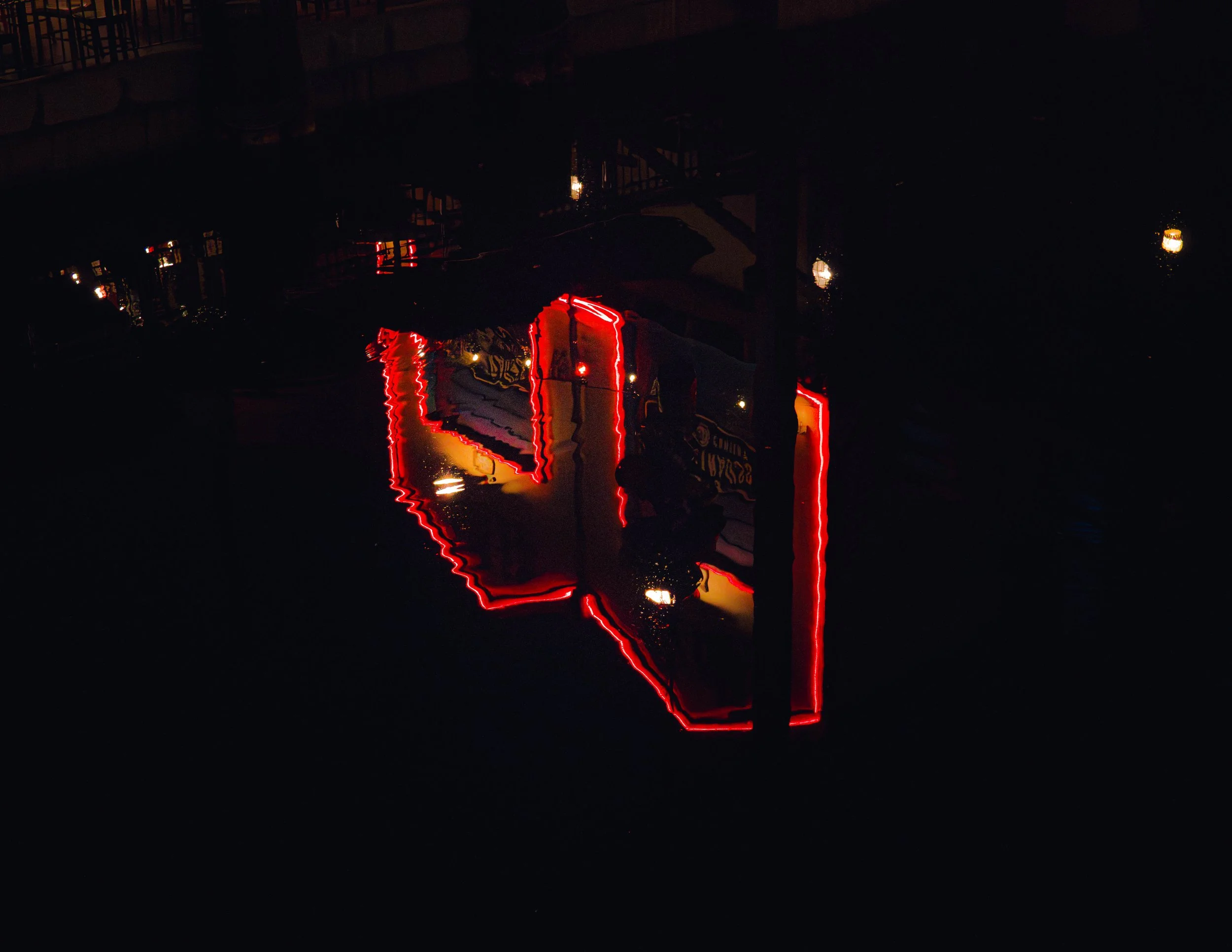 A dark scene with reflections of neon red and yellow lights in a puddle, creating a distorted glowing outline.