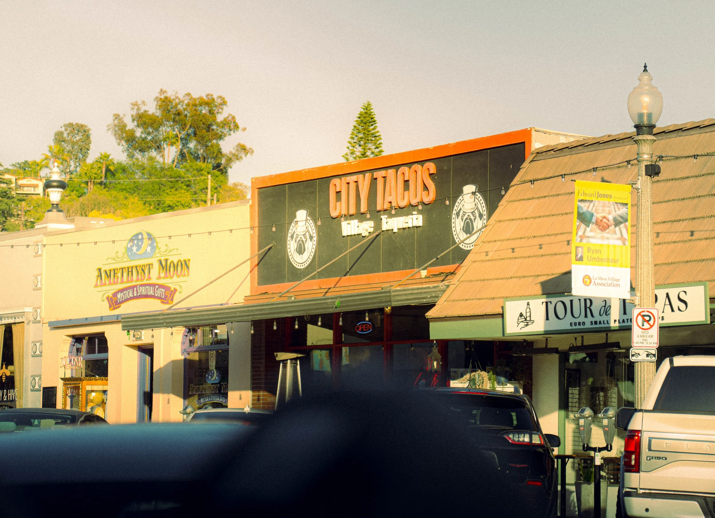 A street scene featuring storefronts with signs, including one for 'City Tacos' and another for 'Amethyst Moon,' with cars parked in front and decorative street lamps.