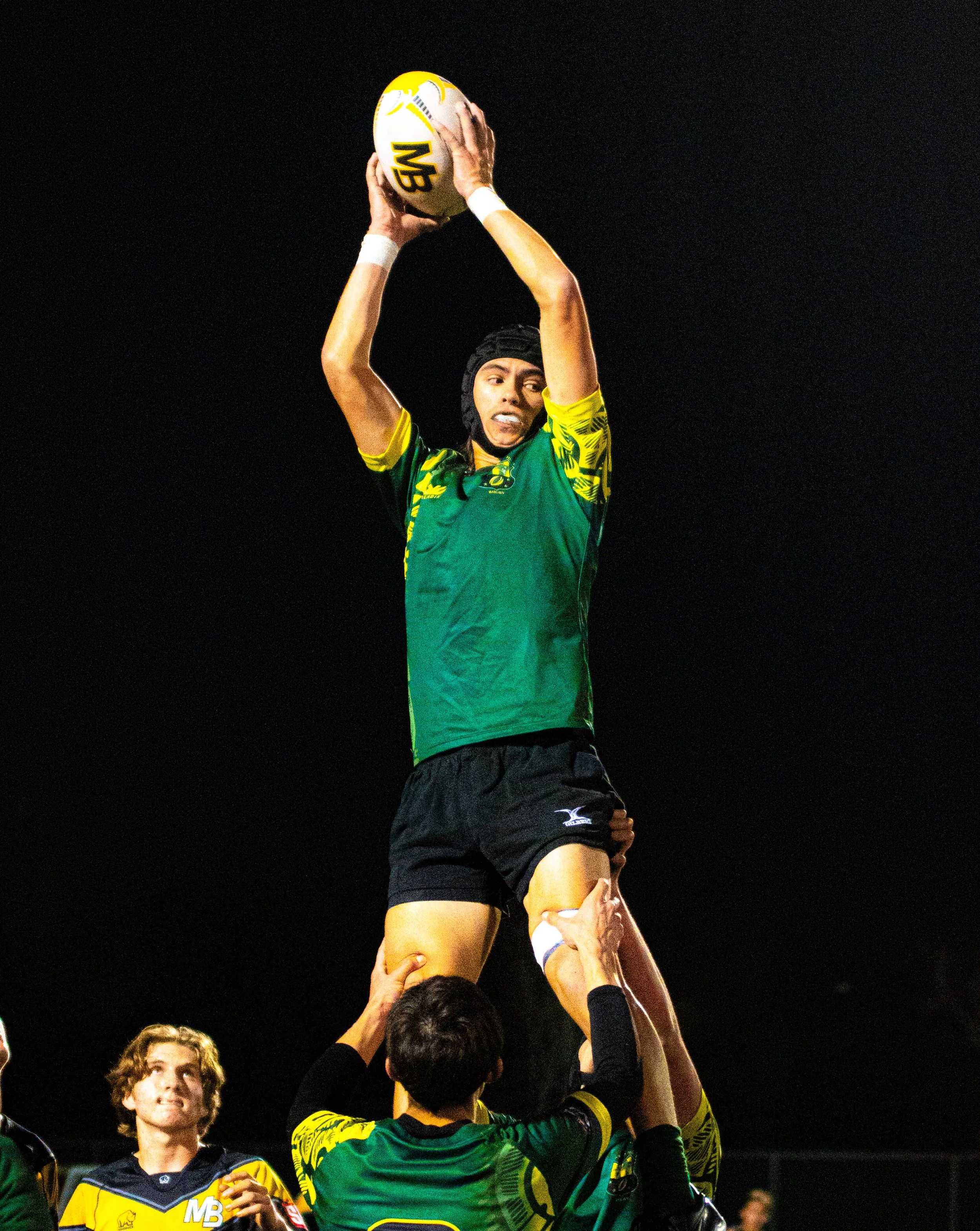 Rugby players perform a lineout lift, with one player in green holding a ball overhead while teammates lift him, at night under stadium lights.