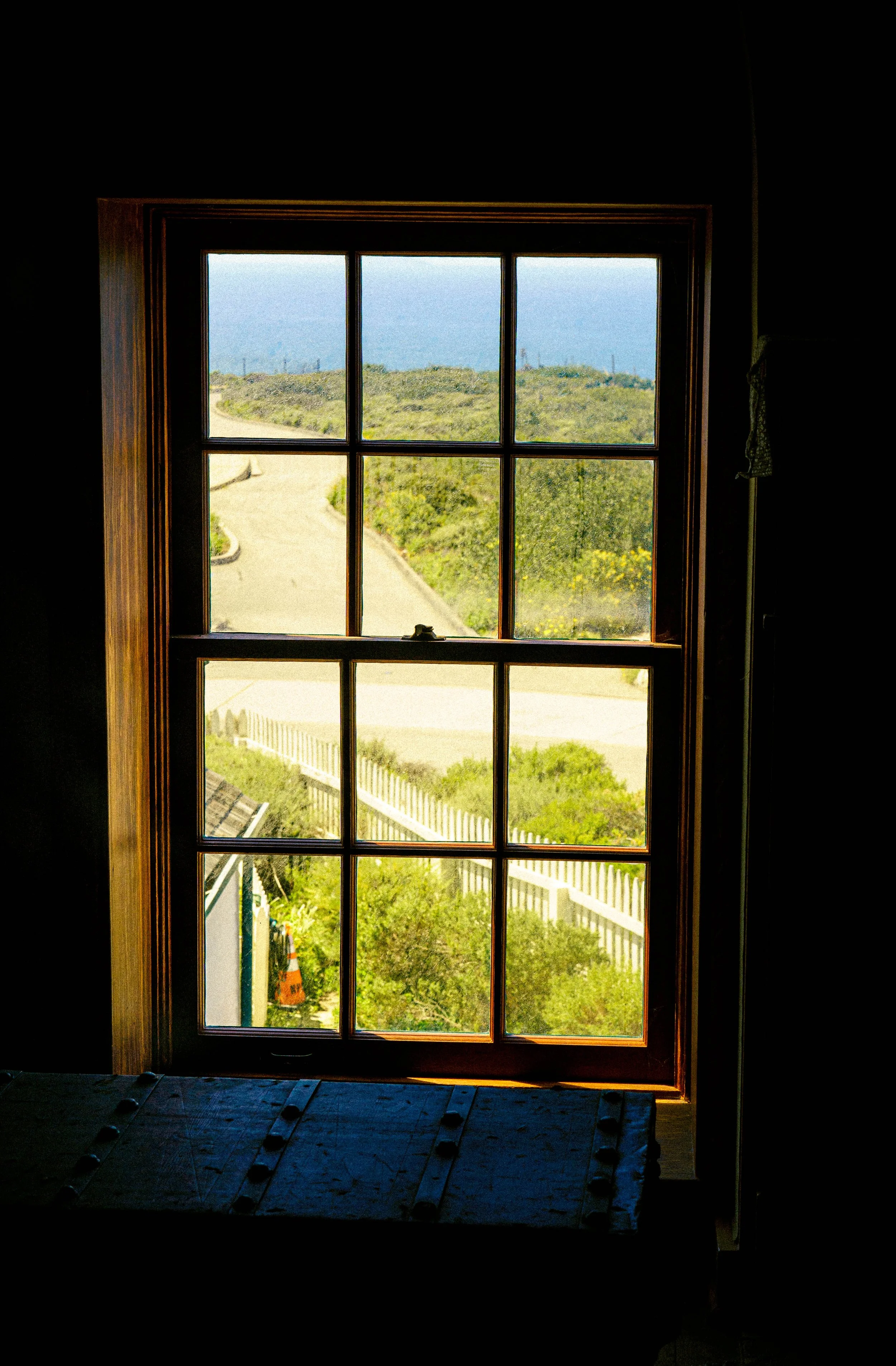 View through a rectangular window with wooden frame showing a landscape with a dirt path, green bushes, and a distant horizon with blue sky and water.