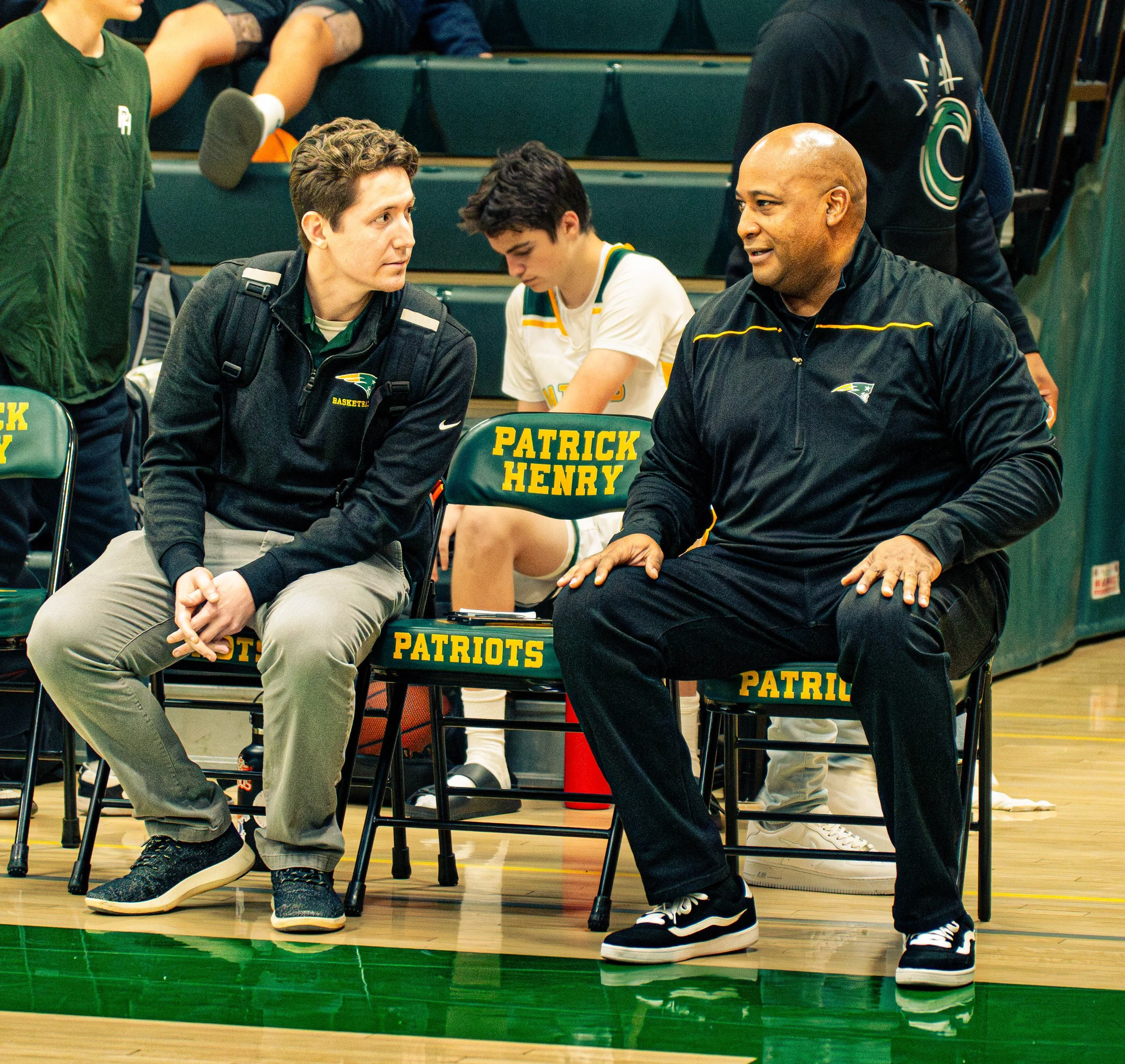 Two young men sitting and talking in a basketball arena, with a person in a white basketball uniform sitting behind them. The young man on the left is wearing a black jacket and khaki pants, and the other young man wears a black jacket and black snea