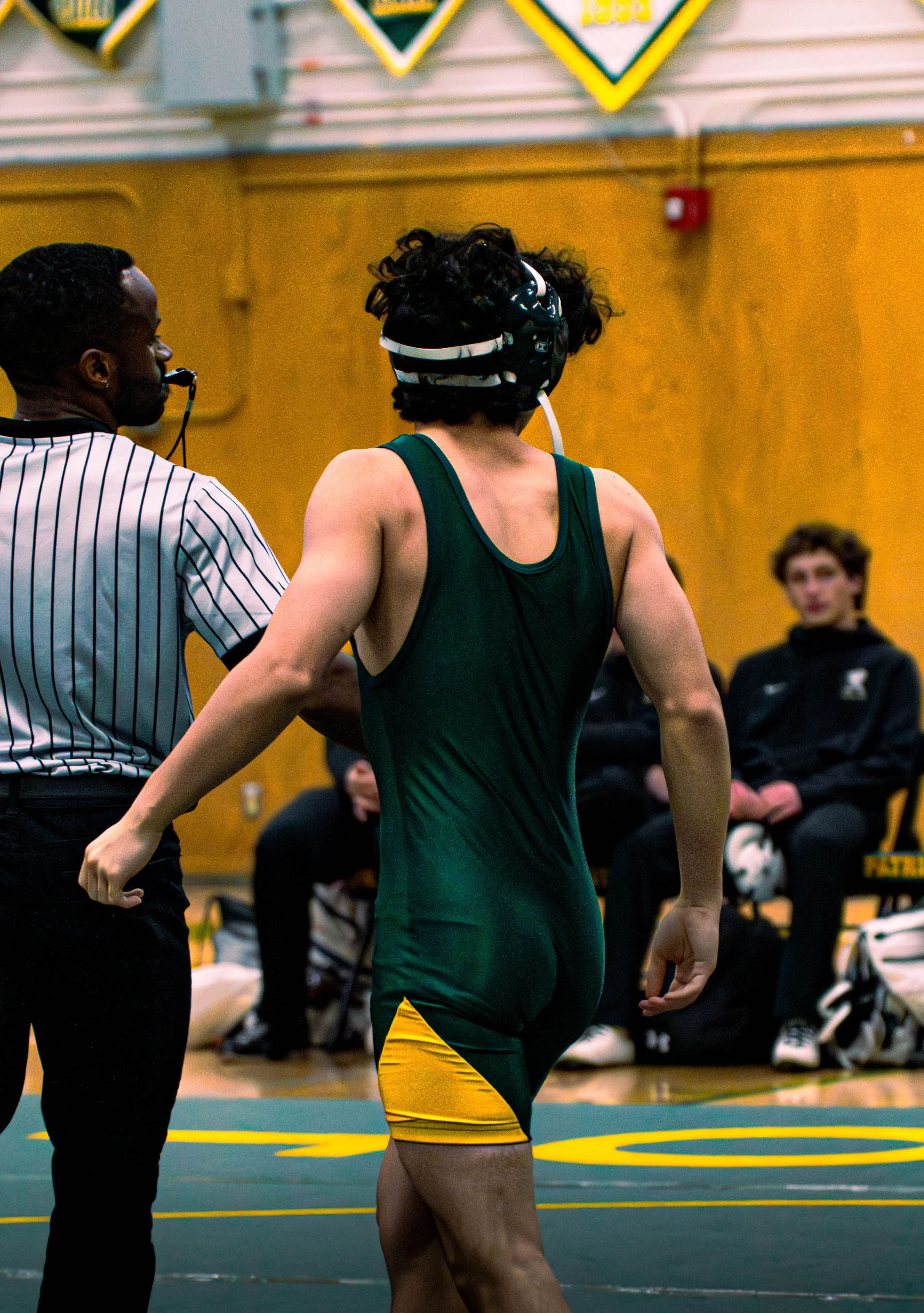 An athlete in a green and yellow uniform with a black protective helmet is standing on a wrestling mat, speaking with a referee in a striped shirt, during a wrestling match in a gymnasium with yellow walls and other people in the background.