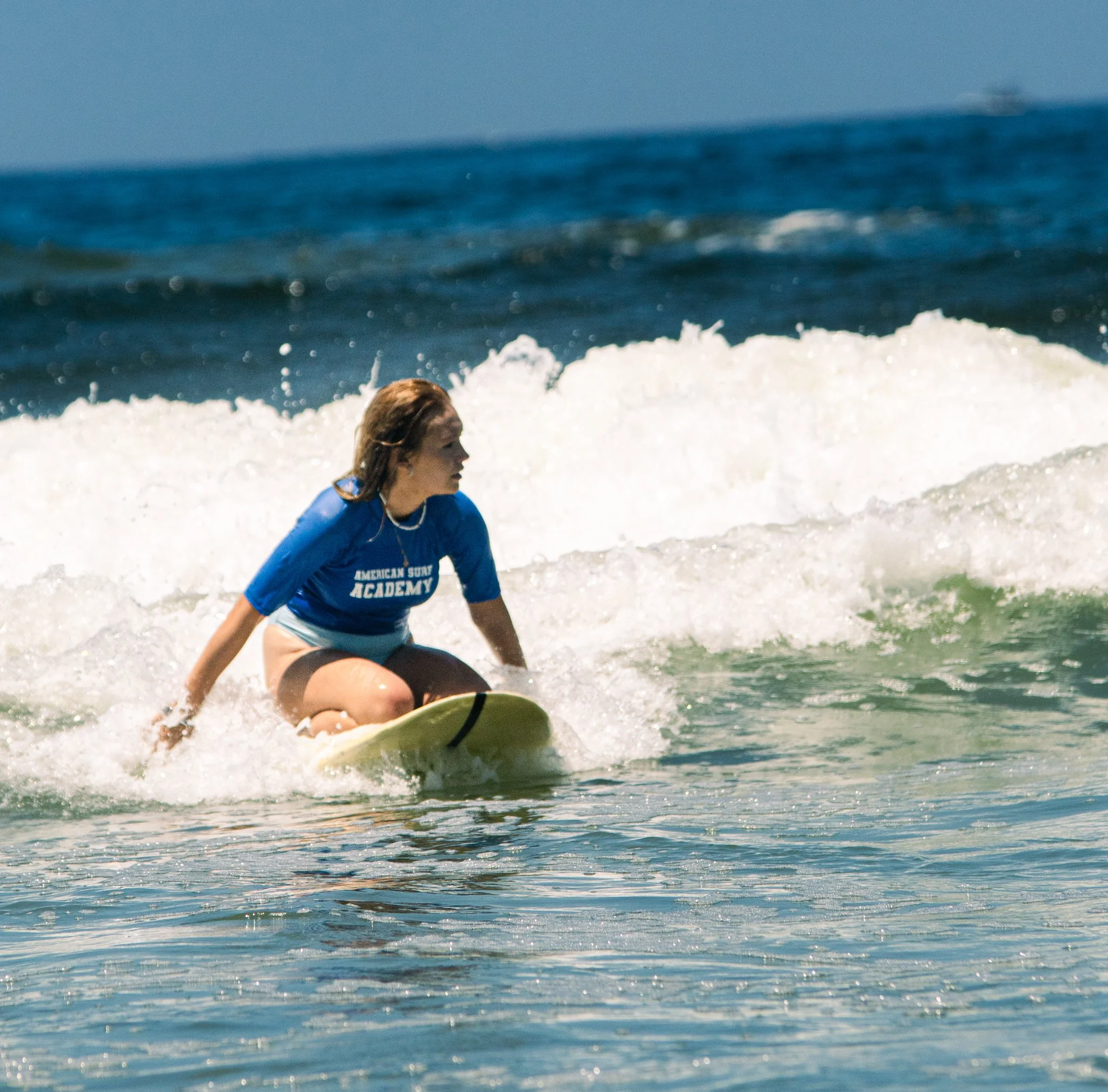 A person wearing a blue shirt crouches on a surfboard in the ocean with waves, under a clear sky.