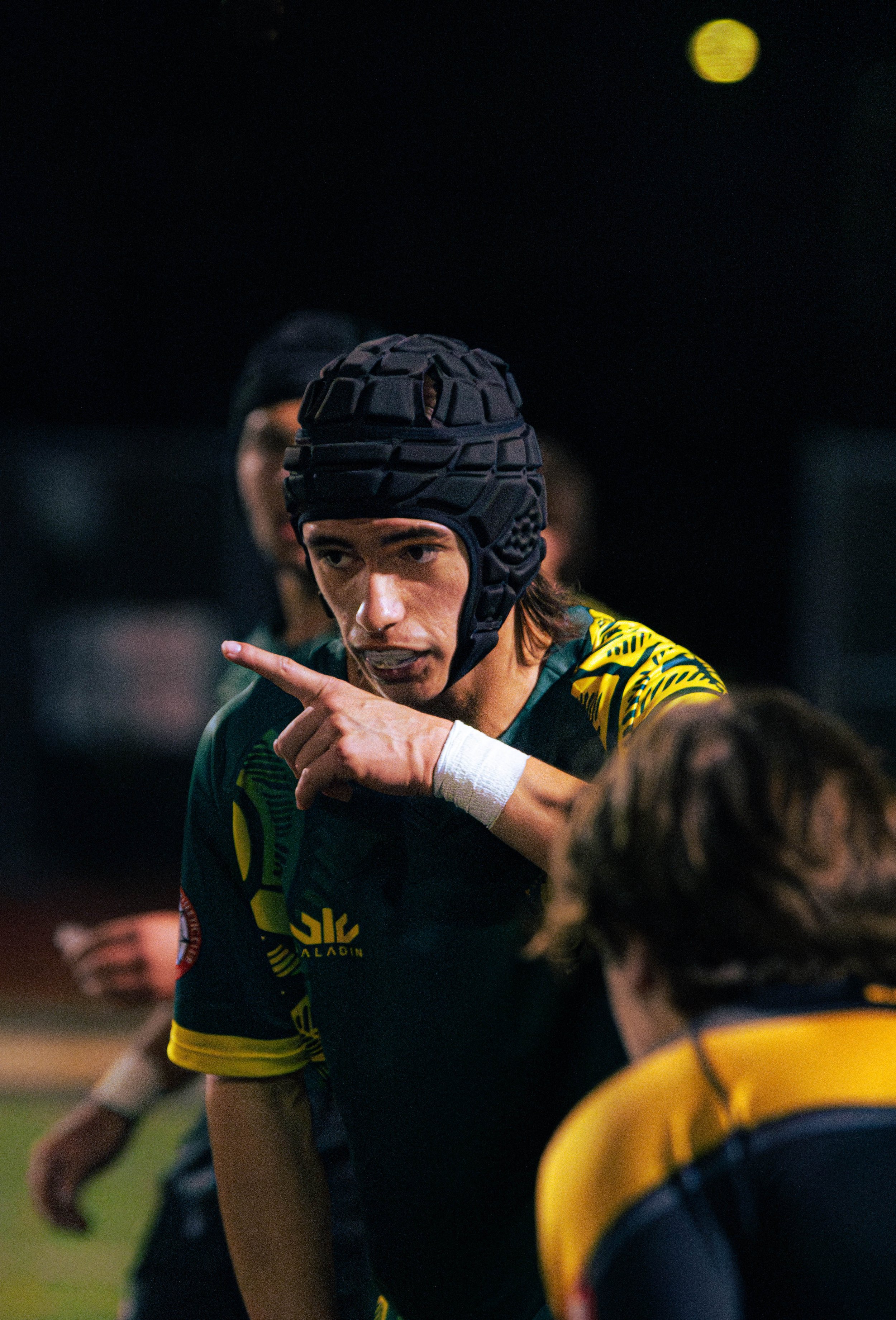 A young male rugby player, wearing a black scrum cap and team jersey, is in a huddle during a game, pointing and talking to teammates on the field at night under stadium lighting.