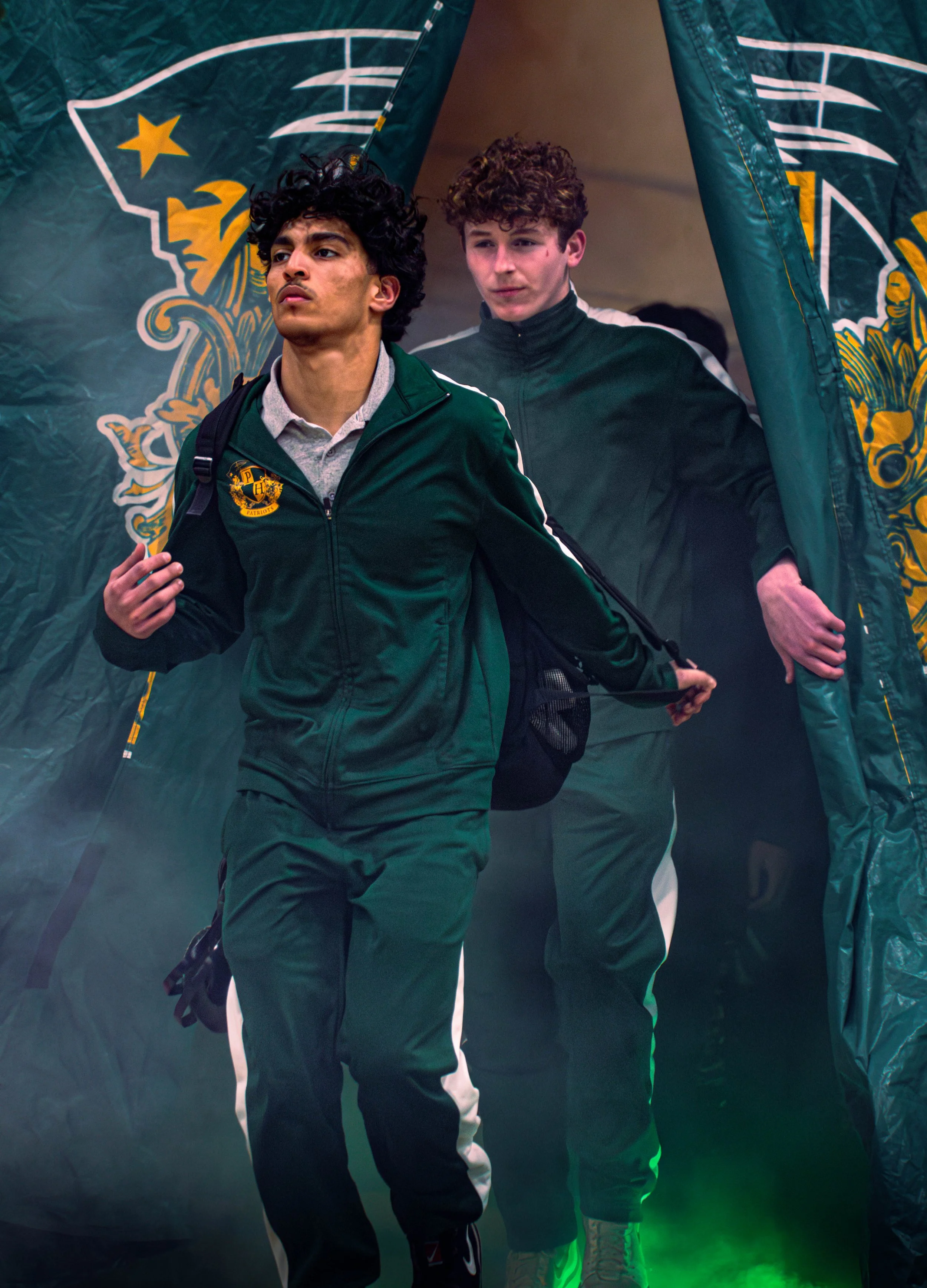 Two young men walking out of a green and yellow Patriots-themed tunnel at a sports event.