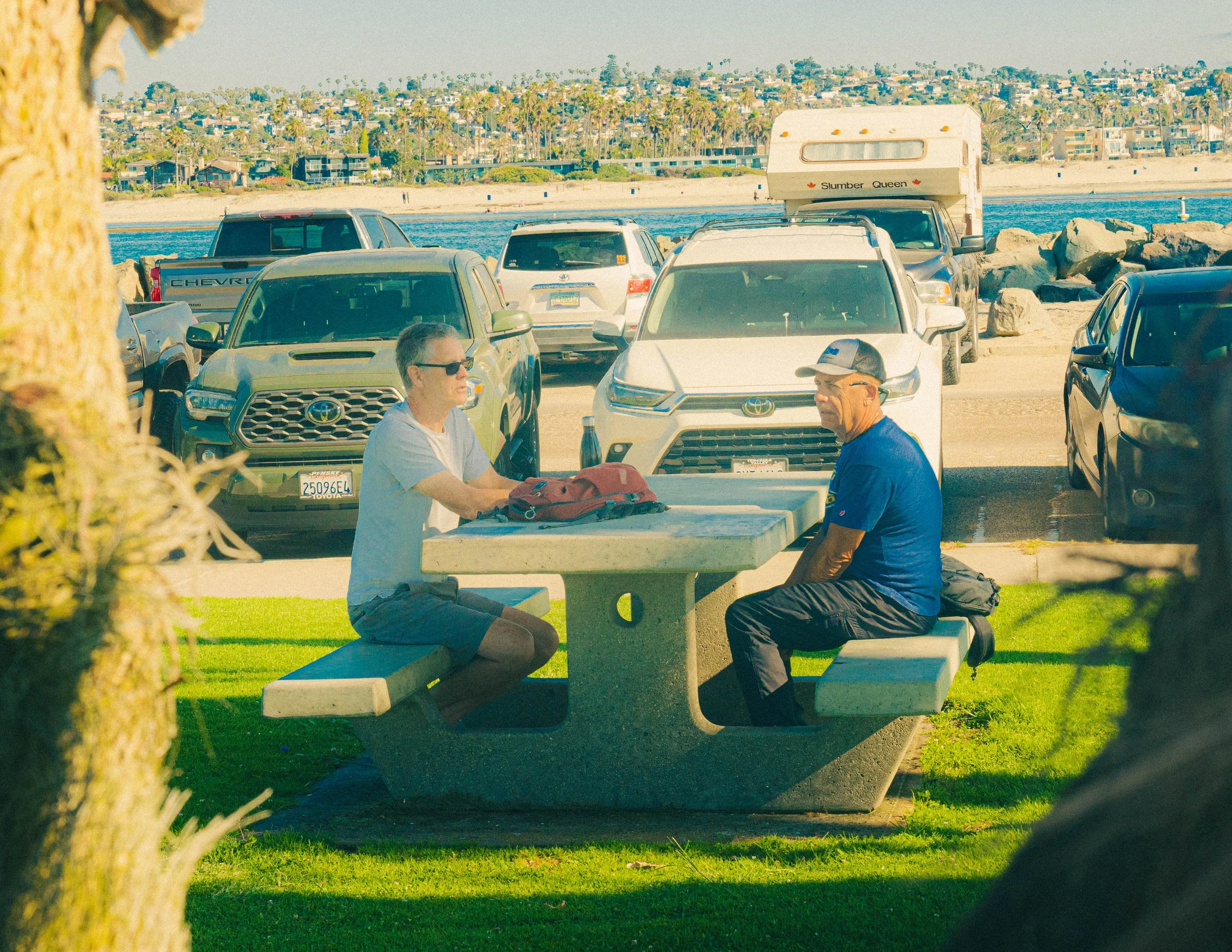 Two men sitting on benches near a parking lot by the beach, with cars and a camper in the background, during daytime in a coastal area with palm trees and houses visible across the water.
