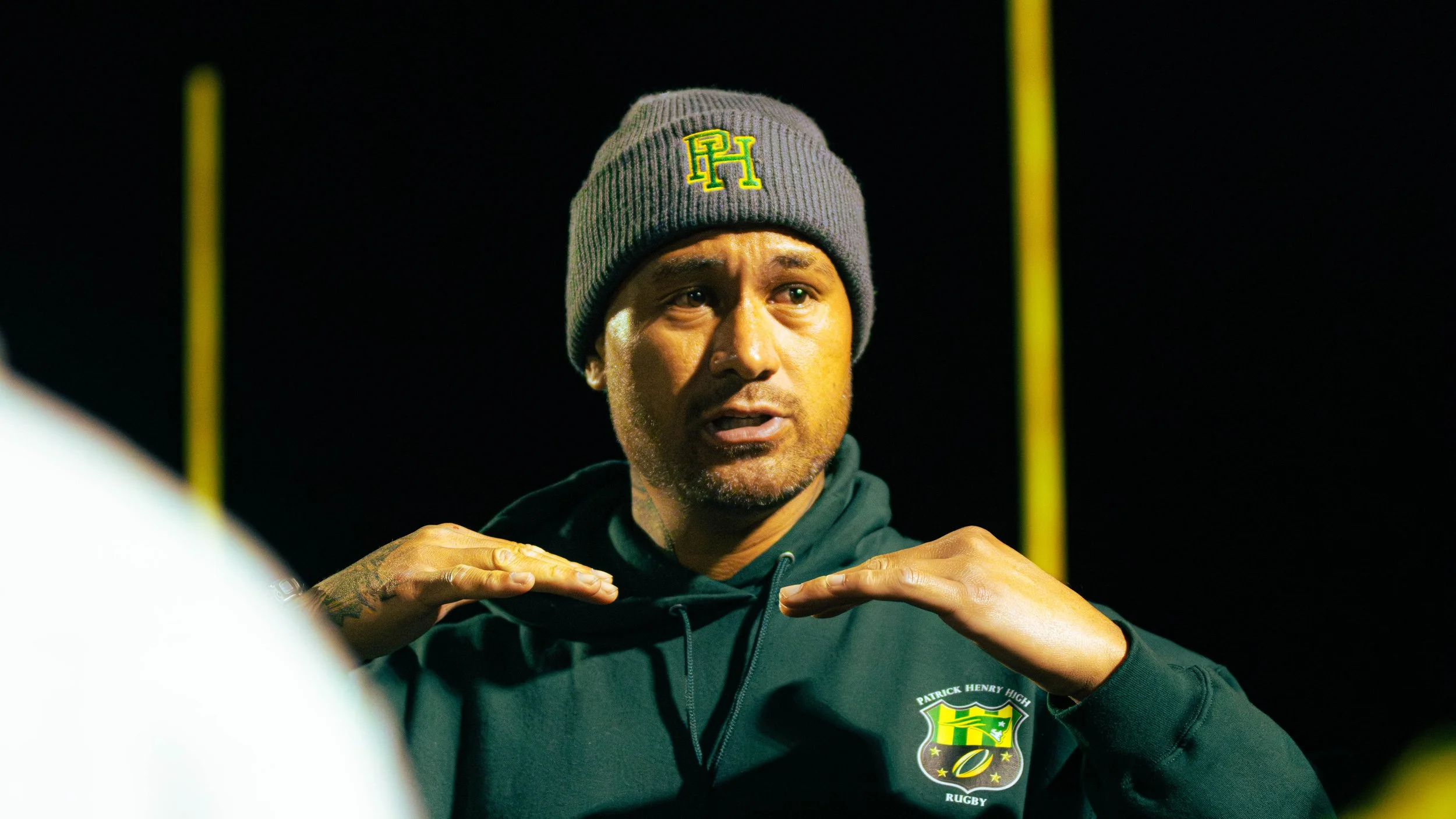 A man wearing a gray knit beanie with yellow and green logo and a black hoodie with a crest and the word 'RUGBY', standing outdoors at night with yellow goal post in the background.
