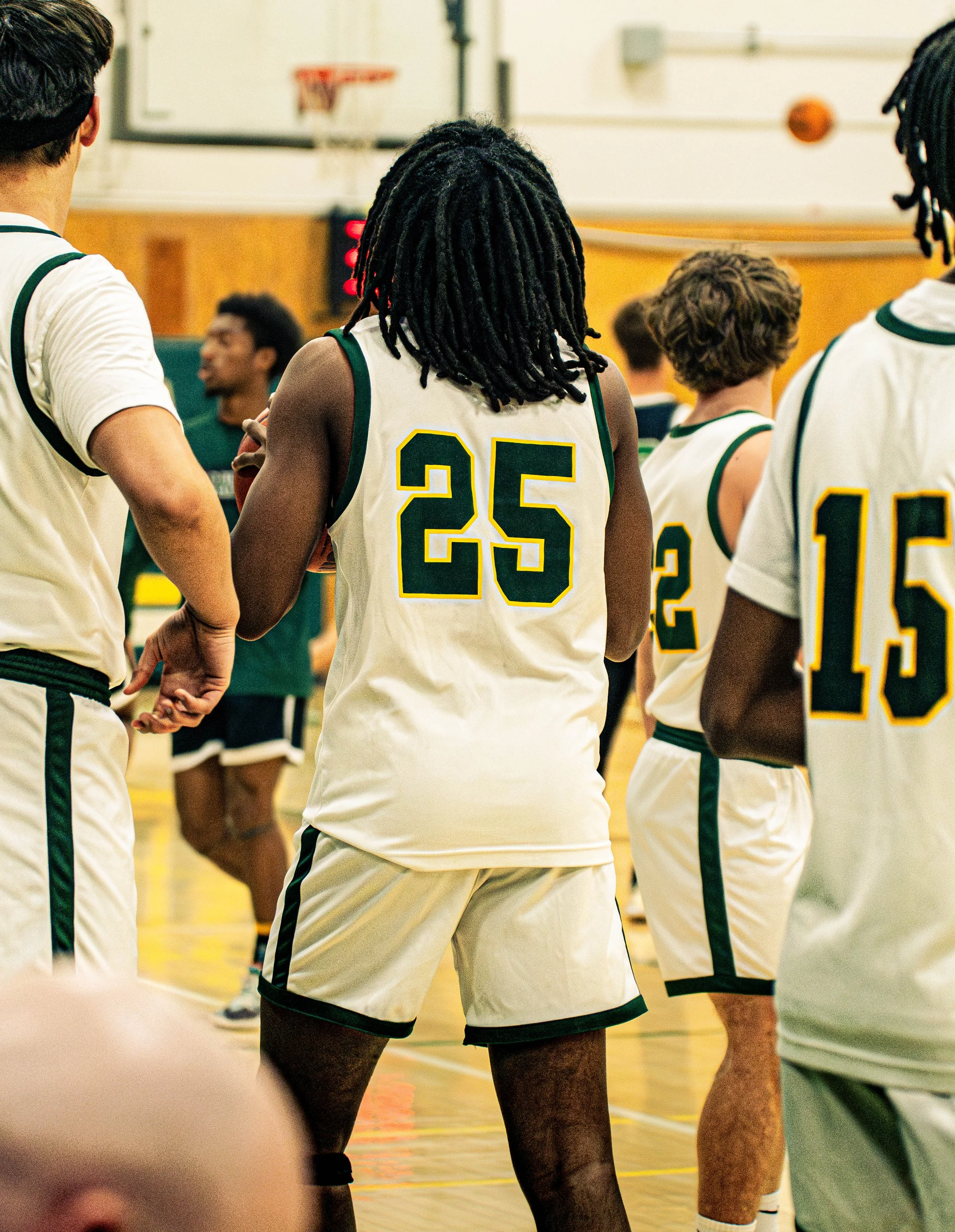 A group of basketball players in white and green uniforms standing in a gymnasium, facing away from the camera, with a basketball hoop visible in the background.