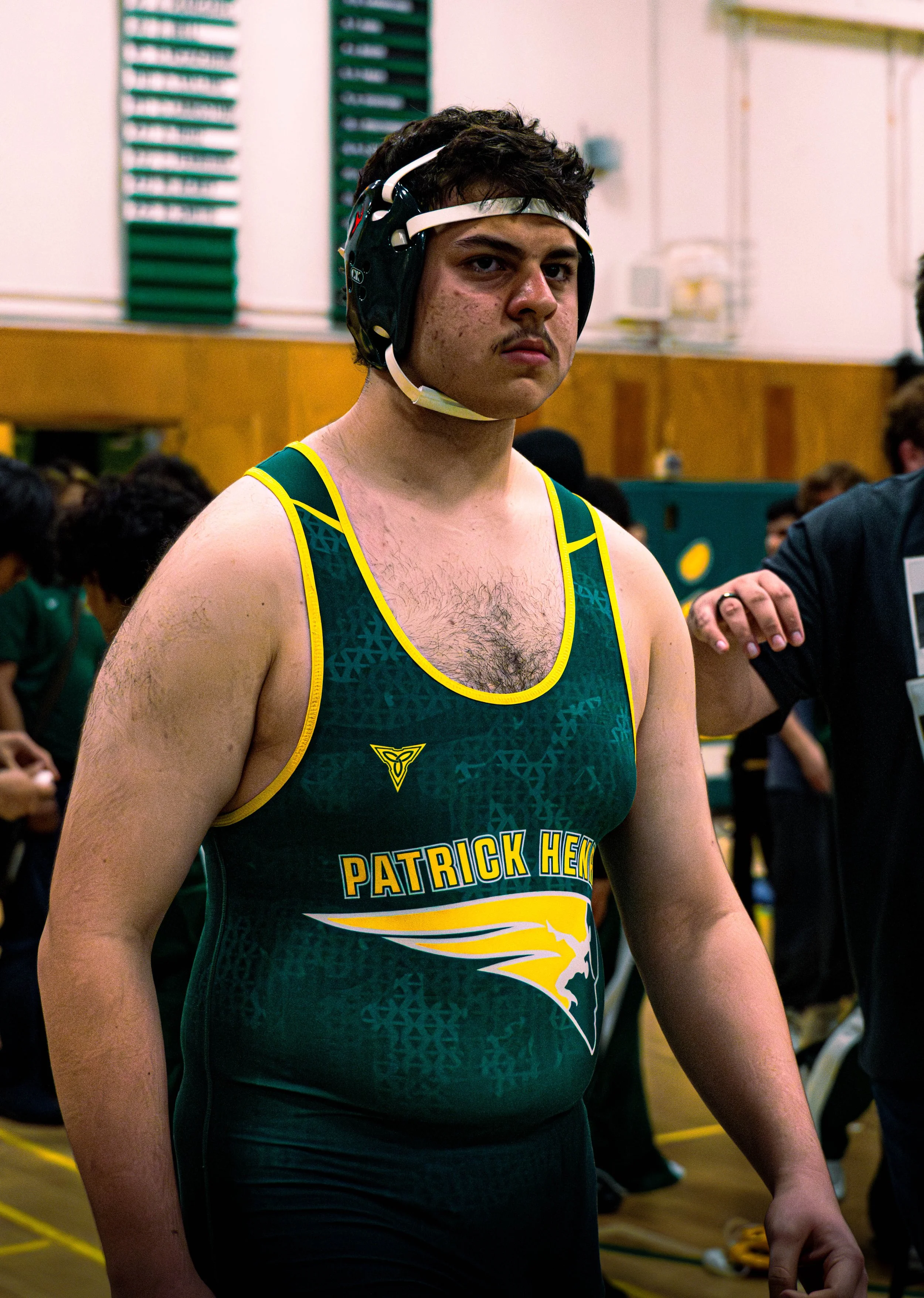 A young male athlete wearing a green and yellow sports singlet with the words "Patrick Hen" and a kangaroo logo on it, standing indoors in what appears to be a sporting event setting, with a focused expression and wearing a headgear.