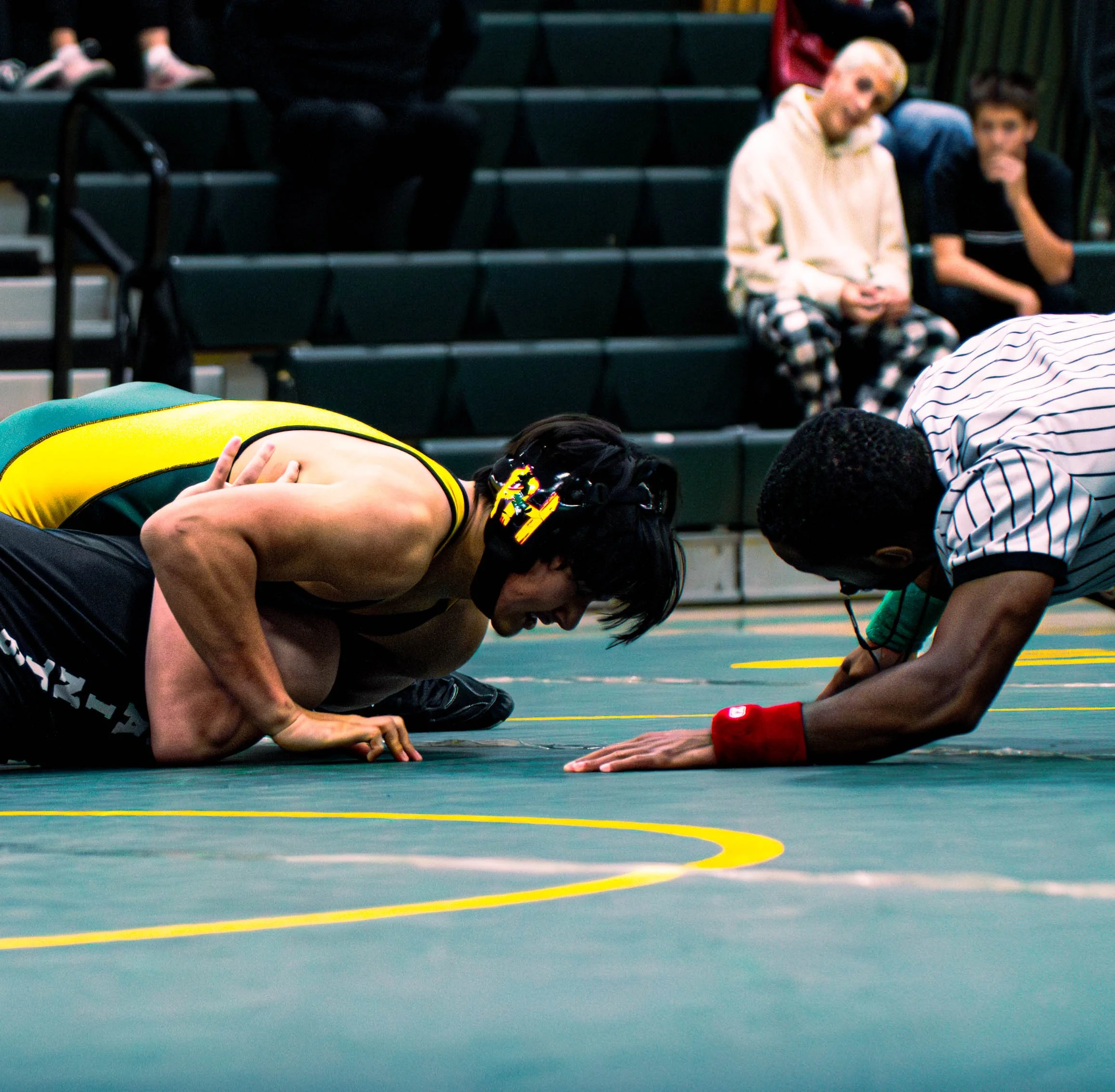 People resting on their knees and lying on the floor in a gymnasium, with some seated on bleachers in the background.
