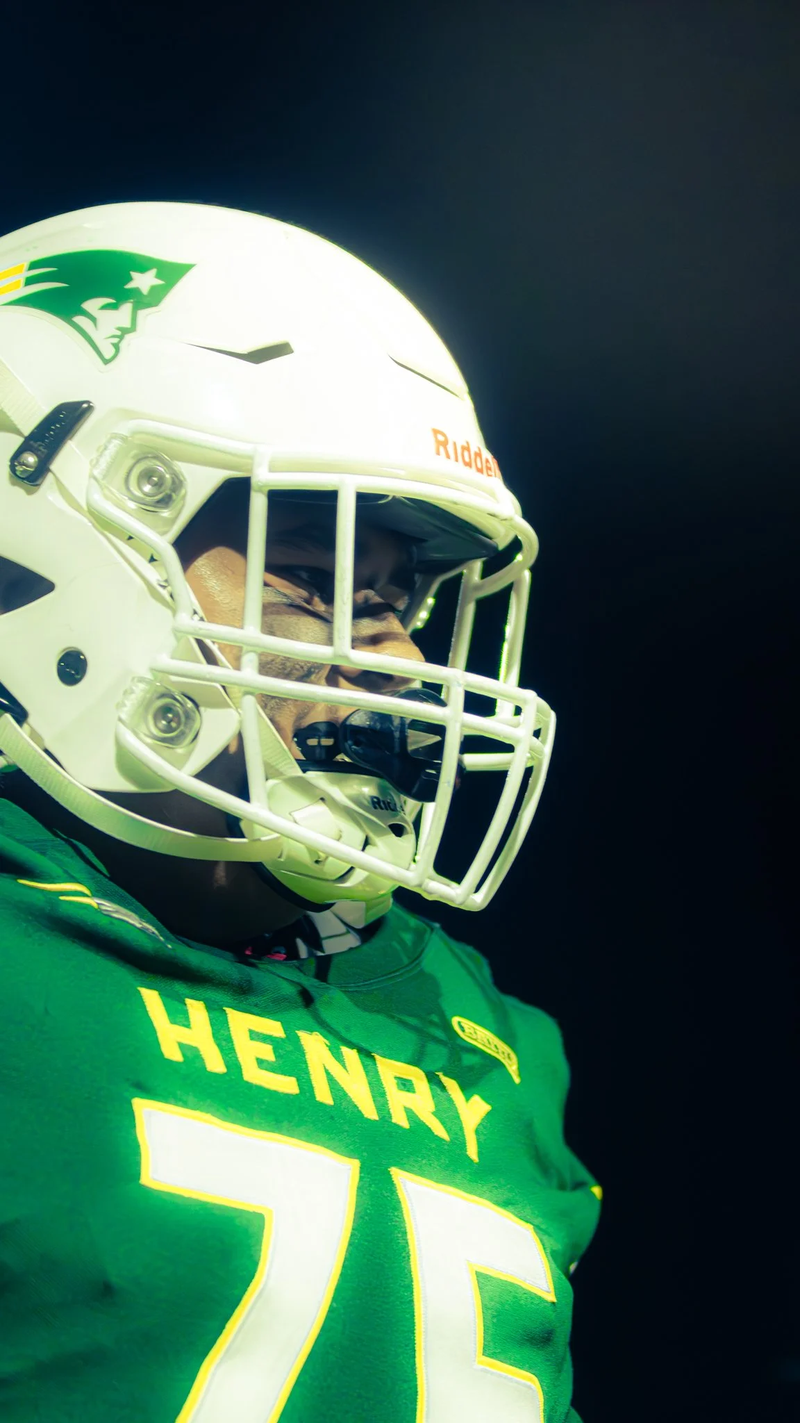 Close-up of a football player wearing a green jersey with yellow lettering and the name "HENRY" on the chest, a white helmet with a green logo of a pirate, a face shield, and a gray face mask, appearing focused against a dark background.