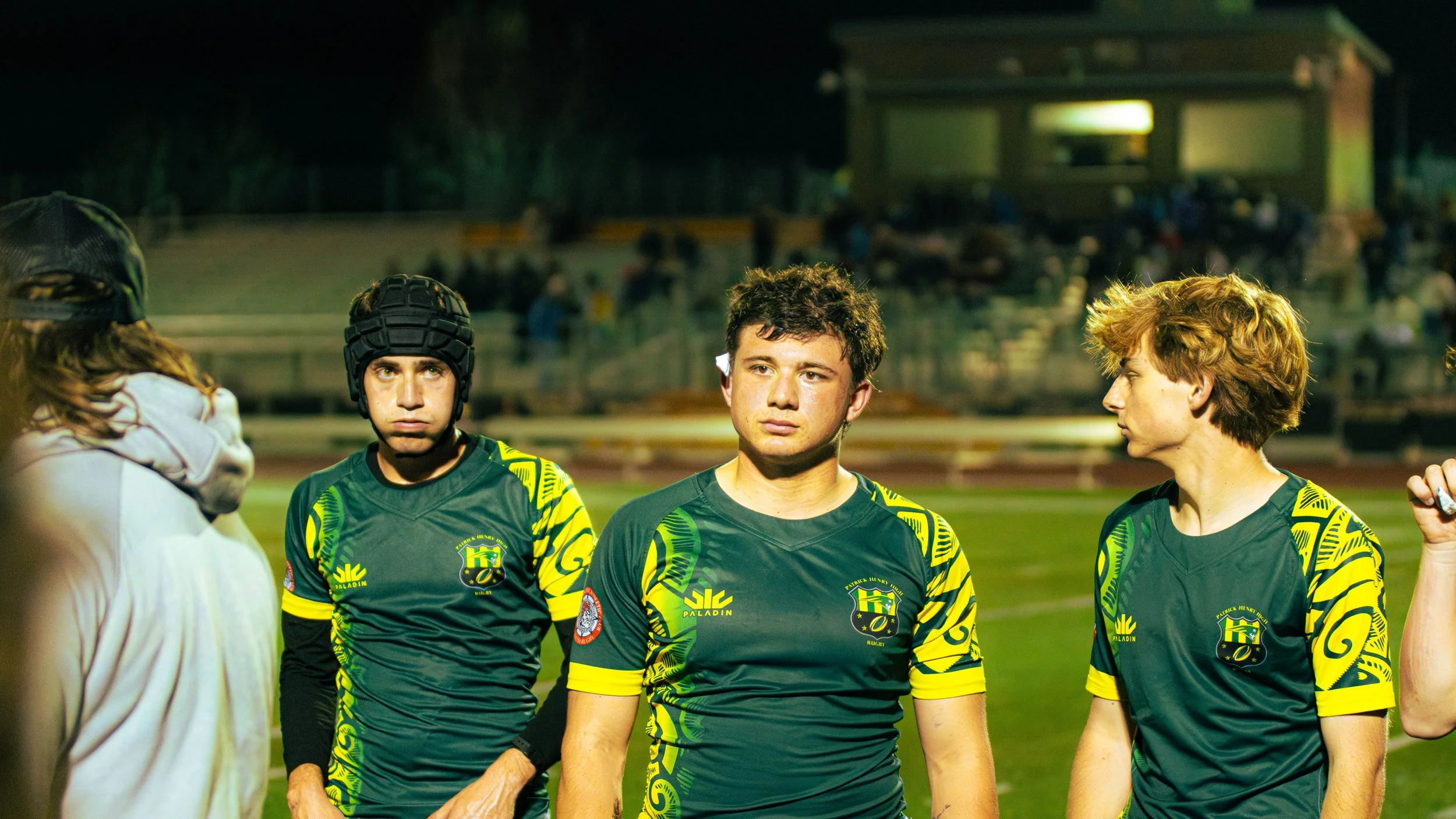 Group of rugby players in green and yellow uniforms standing on a field at night, with a coach or referee talking to them.