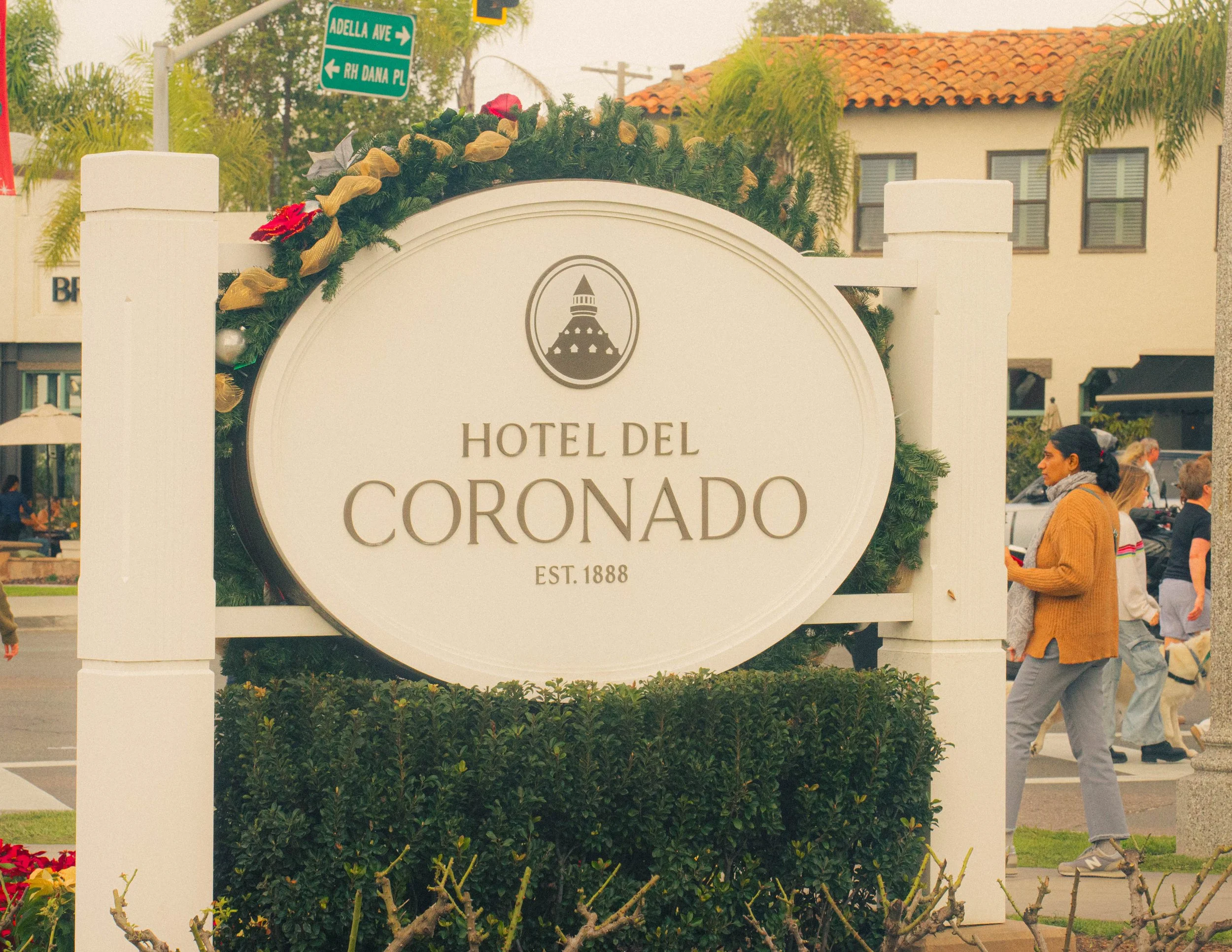 Street sign reading Adella Avenue and RH Dana Place in a downtown area with people walking, lush greenery, and a building with a tiled roof in the background.