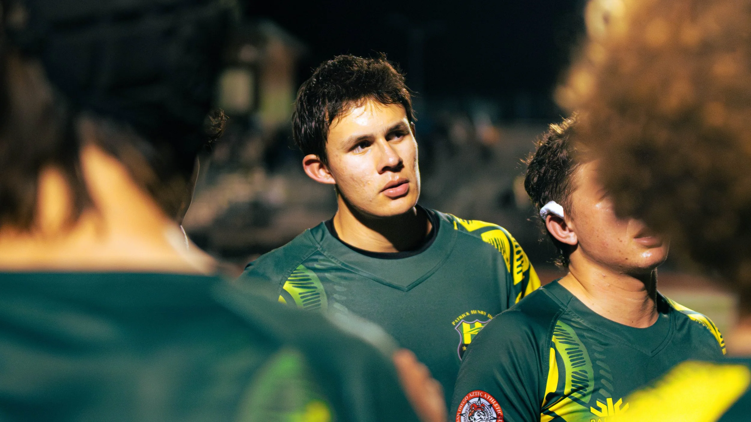 A group of young male soccer players wearing black jerseys with yellow details, standing closely together during a game or practice, with serious expressions and short dark hair.