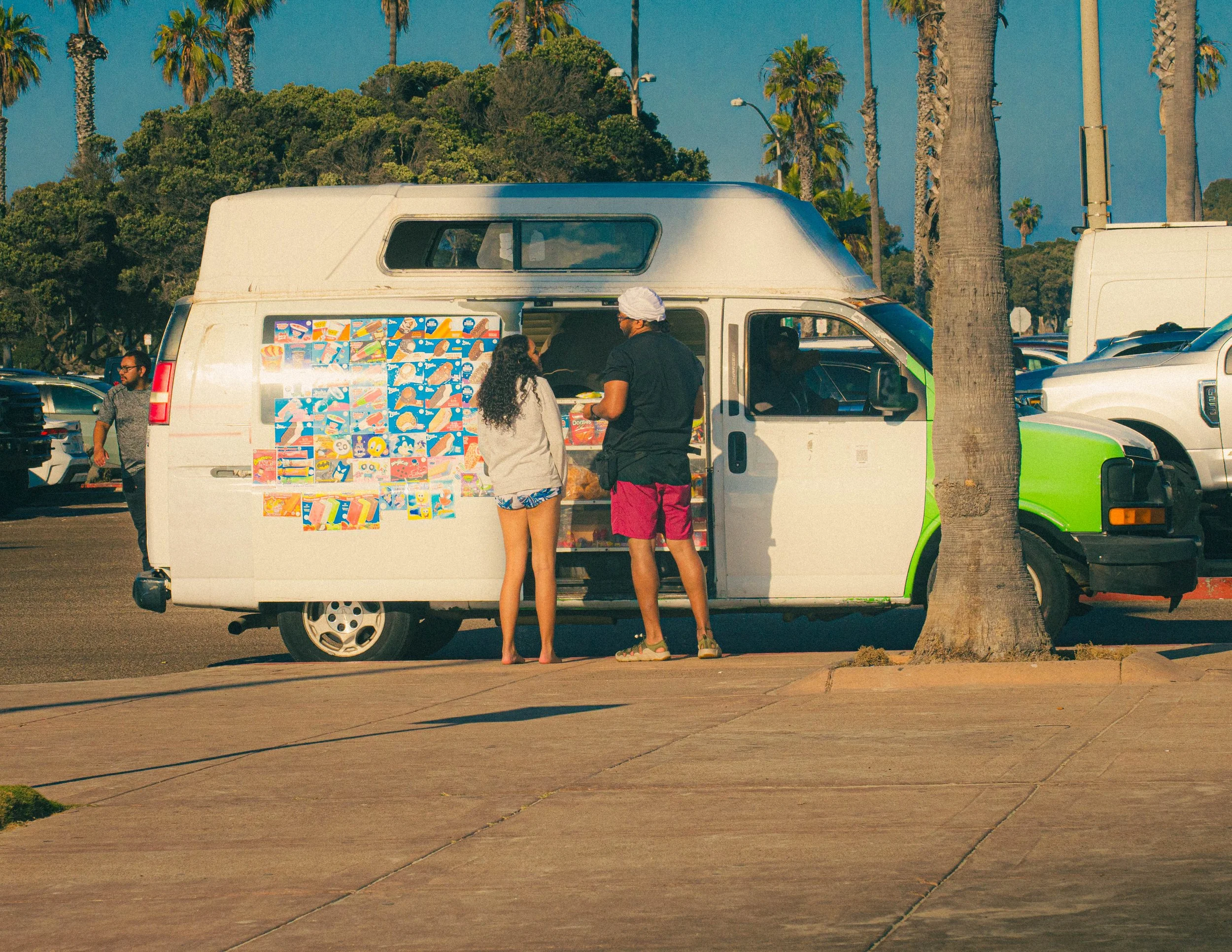 A white food truck with colorful cartoon stickers on its side, parked in a lot with tall palm trees in the background, and people standing nearby. It appears to be late afternoon or early evening.