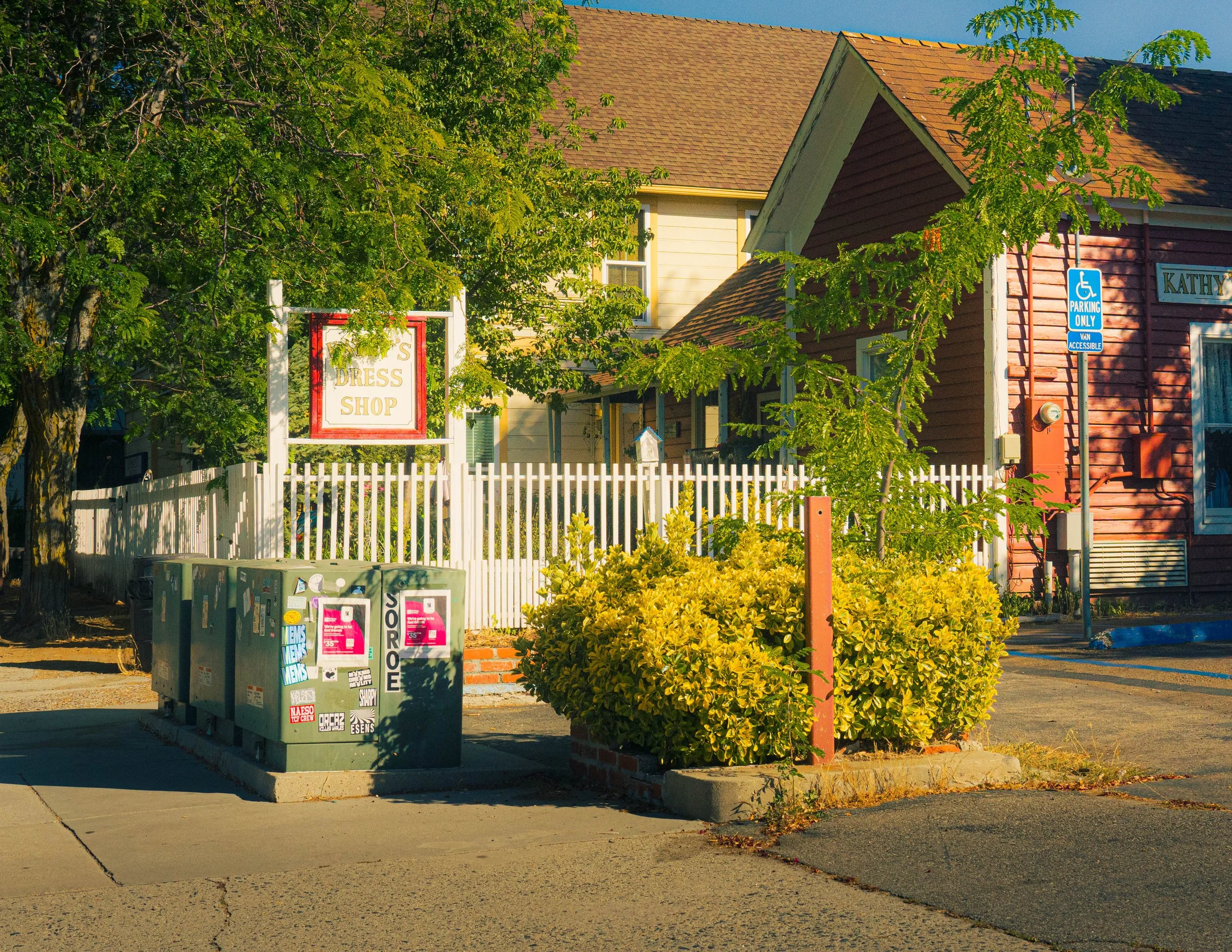 A street view showing a yellow and red house with a brown tiled roof, partially obscured by green trees. A white picket fence borders the street and there are utility boxes on the sidewalk. A sign reads 'Kathy' and a blue handicapped parking sign is 