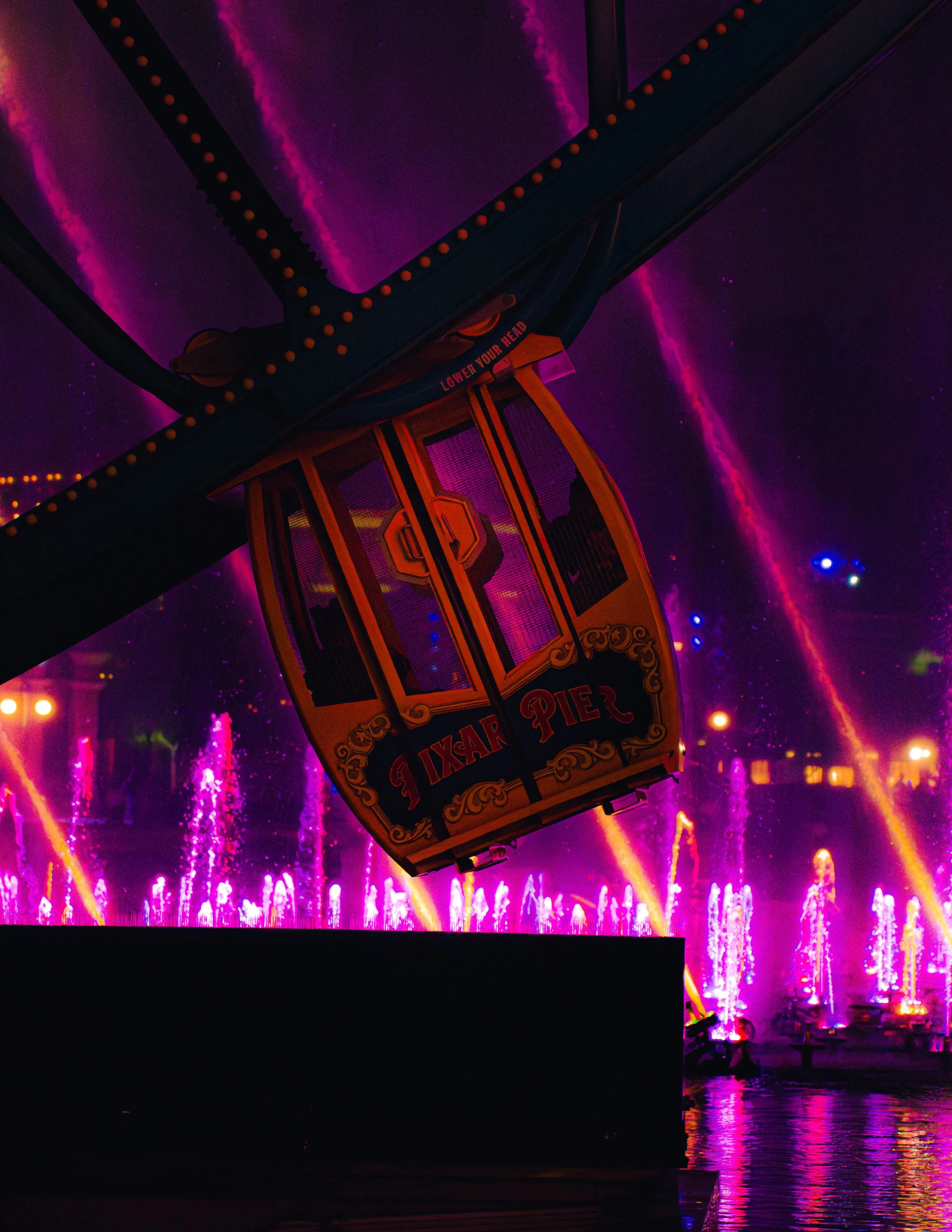 A colorful amusement park ride, possibly a Ferris wheel, illuminated with pink and purple neon lights at night. The background features a laser or light show with streaks of pink, purple, and yellow lights reflecting off water.