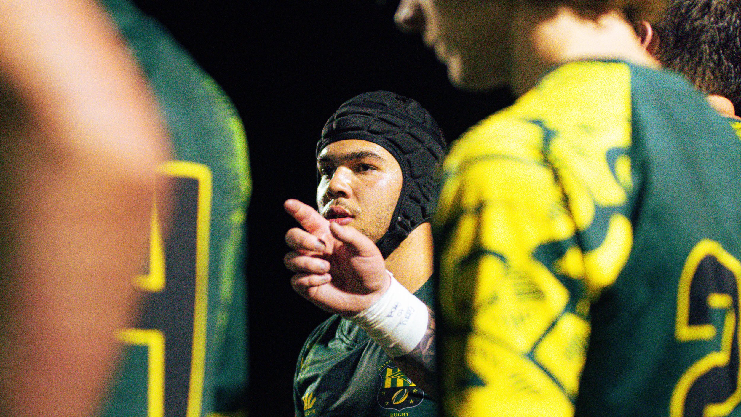 A rugby player wearing a black headgear and black jersey gesturing with their hand while talking to two teammates in yellow and green jerseys.