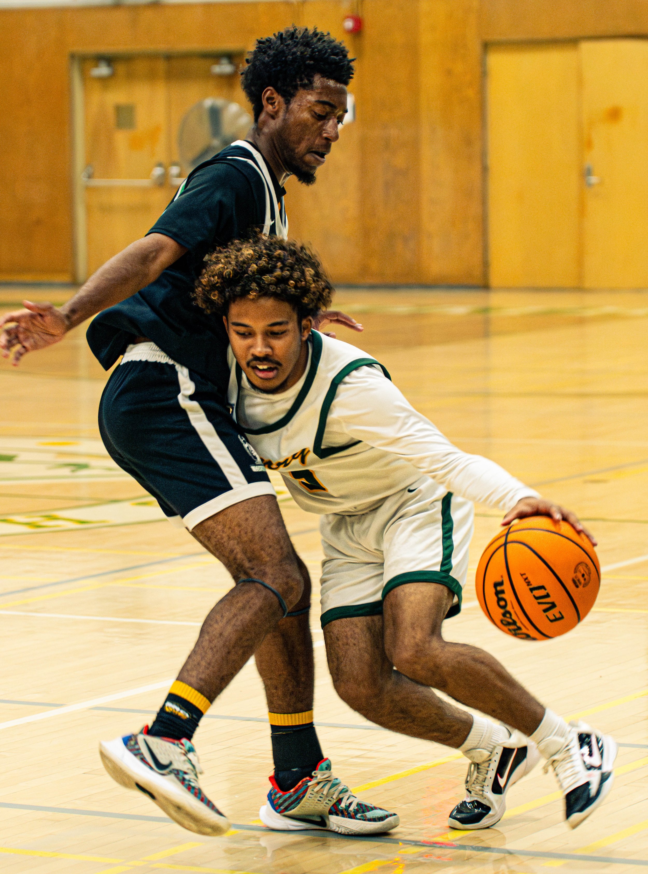 Two male basketball players falling on the court during a game, one in a black jersey and the other in a white jersey with green trim, with a basketball near the white jersey player, in an indoor gymnasium.