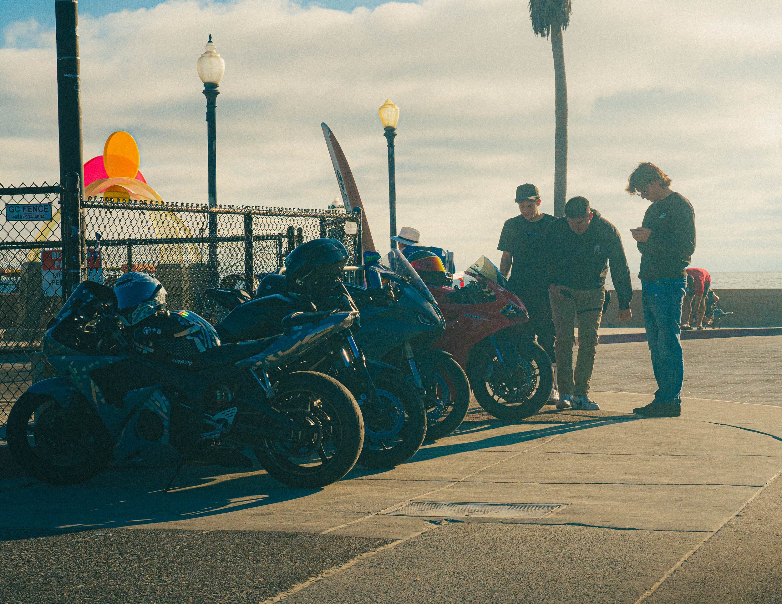 A row of motorbikes parked near a chain-link fence at a beachside location with three people standing next to the bikes. In the background, there are tall lamp posts, a palm tree, and a colorful wind sculpture, with an overcast sky.