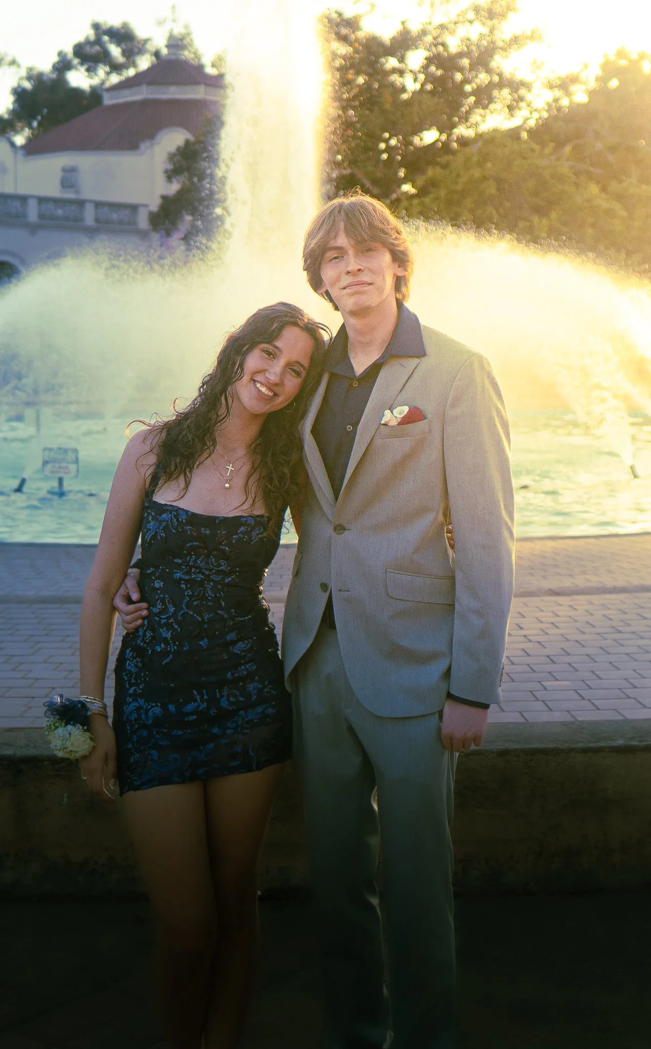 A young woman in a black floral dress with a yellow corsage and a young man in a light gray suit with a dark shirt and pocket square, standing together in front of a fountain with water spraying, during sunset.