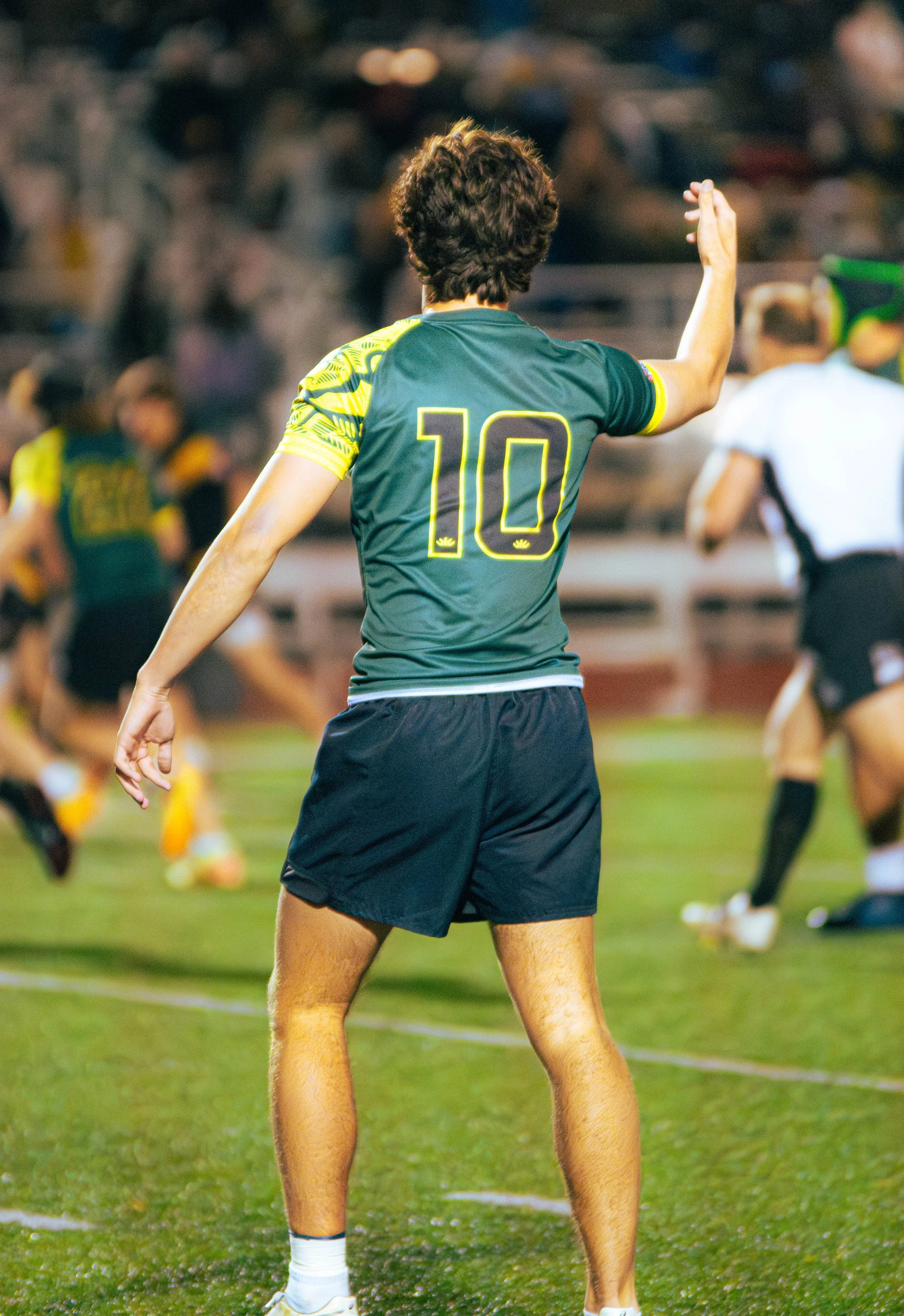 A female soccer player wearing a green jersey with the number 10 on the back, standing on a soccer field during a game. She has short, dark hair and appears to be signaling or gesturing to teammates. In the background, other players and a referee can