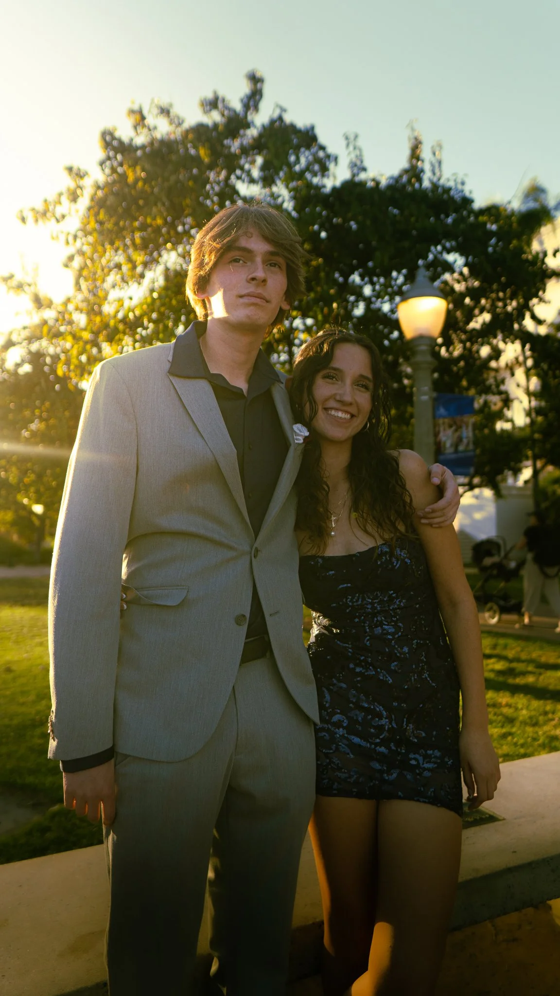 A young man in a light gray suit with a black shirt standing next to a young woman in a black dress with a floral pattern, both smiling, outdoors in the evening with trees, a streetlamp, and people in the background.