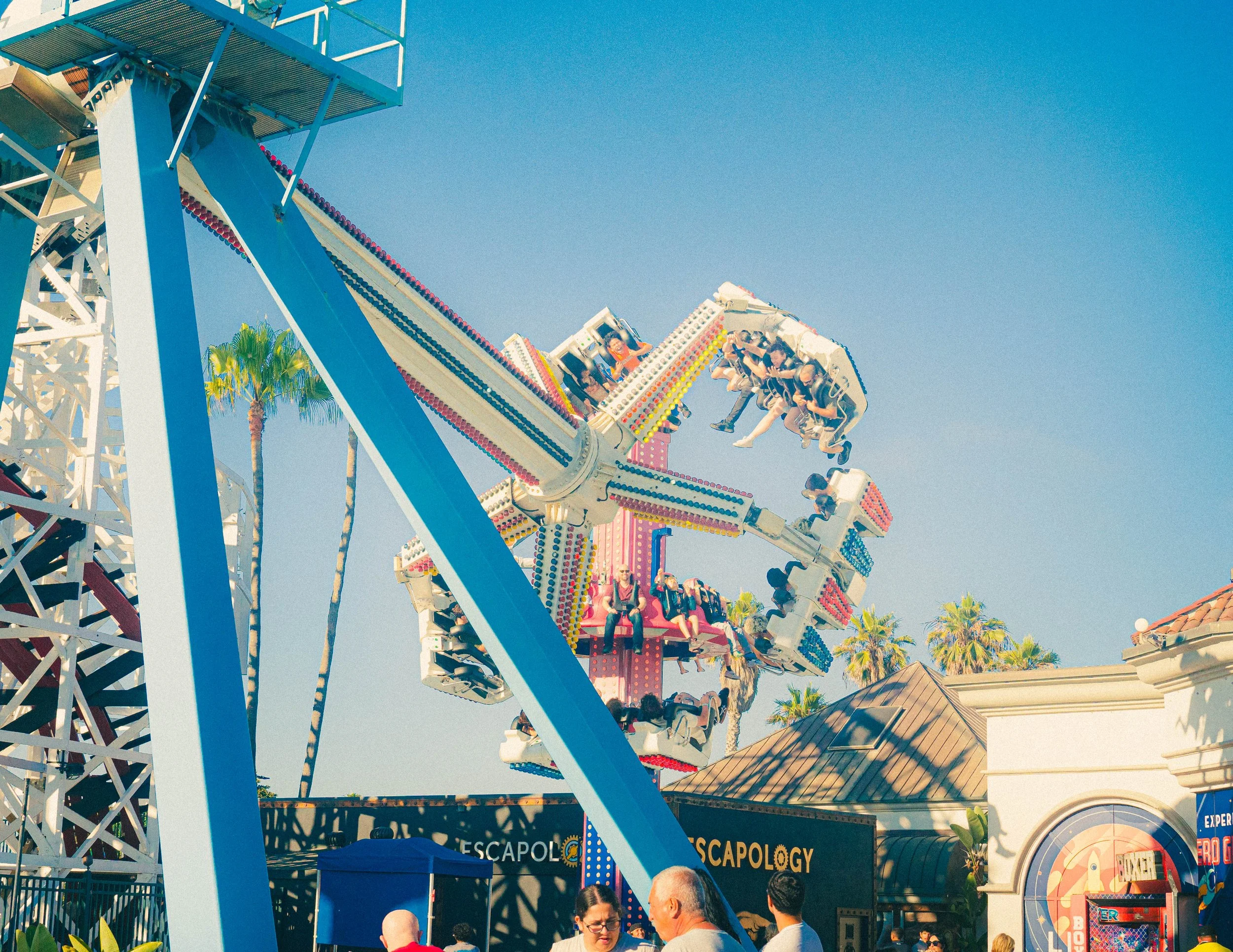 A colorful amusement park ride with people enjoying the ride against a clear blue sky. There are palm trees and a building in the background.