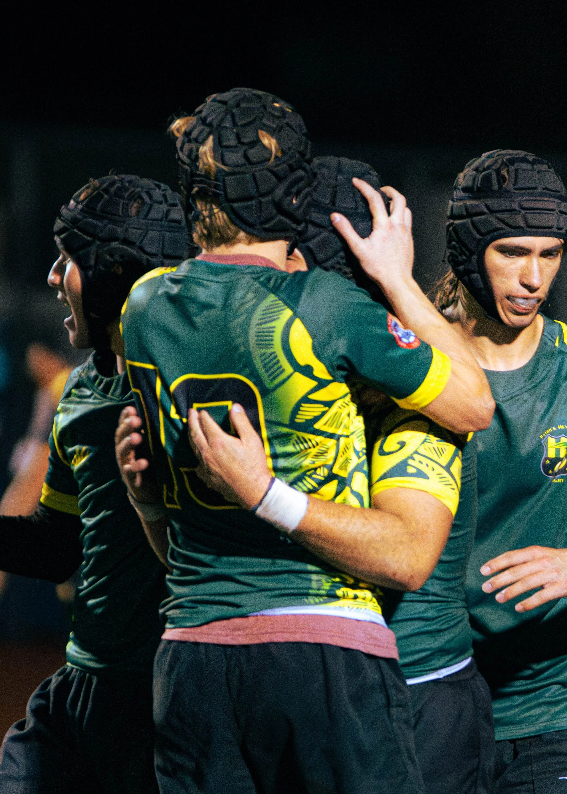 Group of female rugby players in black and yellow uniforms celebrating on the field, wearing protective headgear, embracing each other.