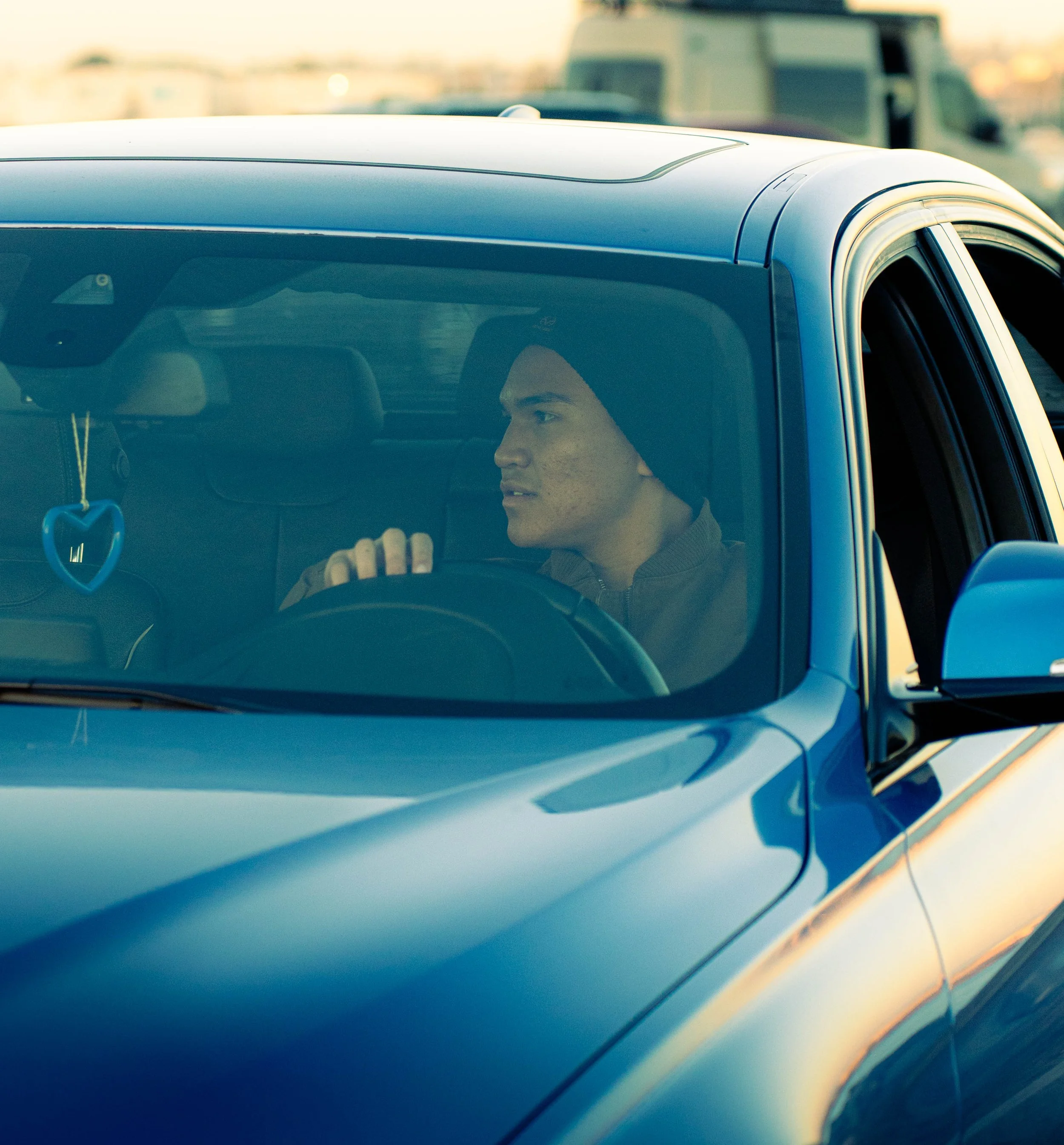 Close-up of a blue car's roof and window with a focus on the driver's side. A person is visible inside, looking to the right with headlights of other vehicles in the background.