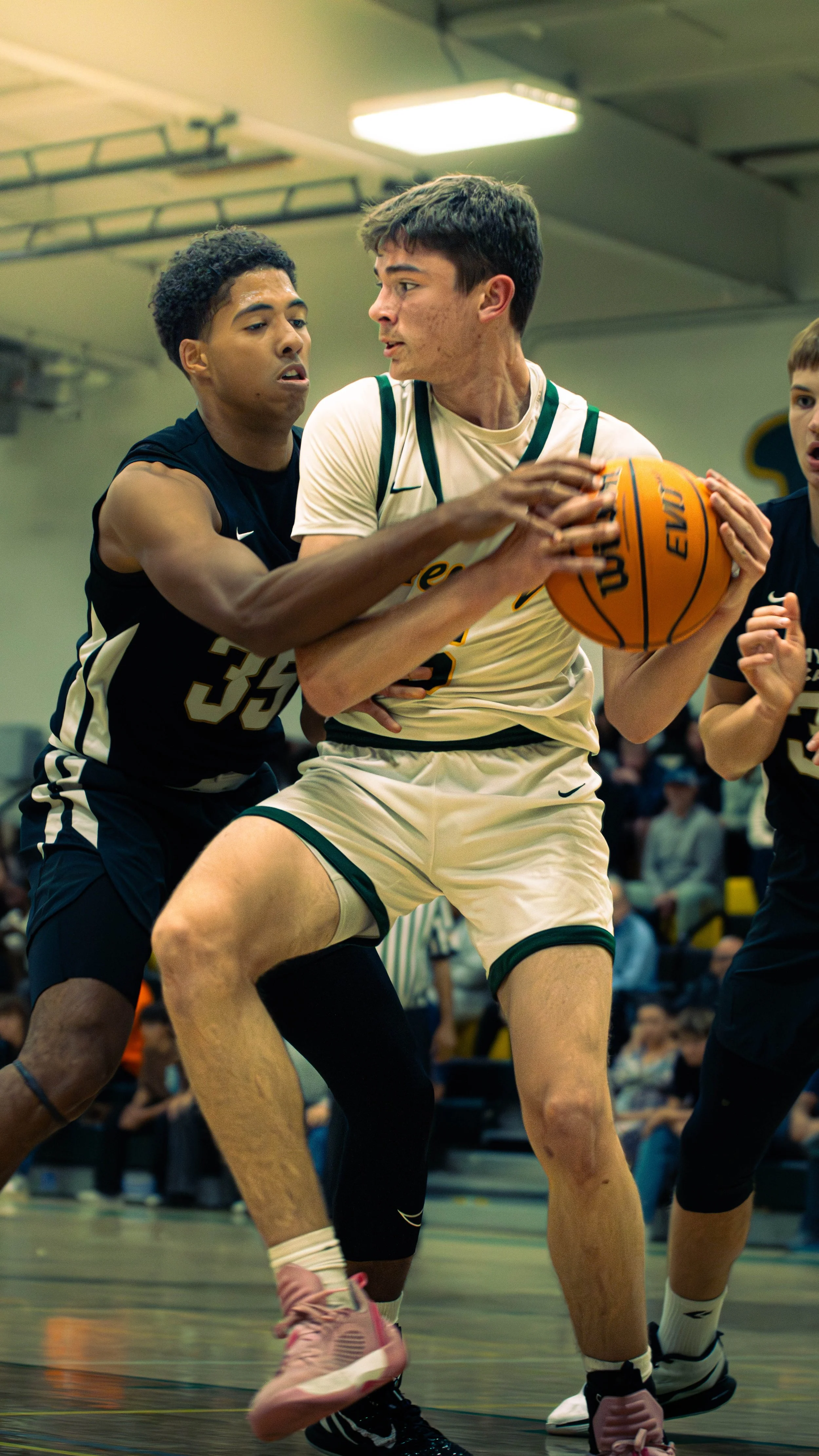Two basketball players in a game, one in a black jersey and one in a white jersey, fighting for possession of the ball inside a gymnasium.
