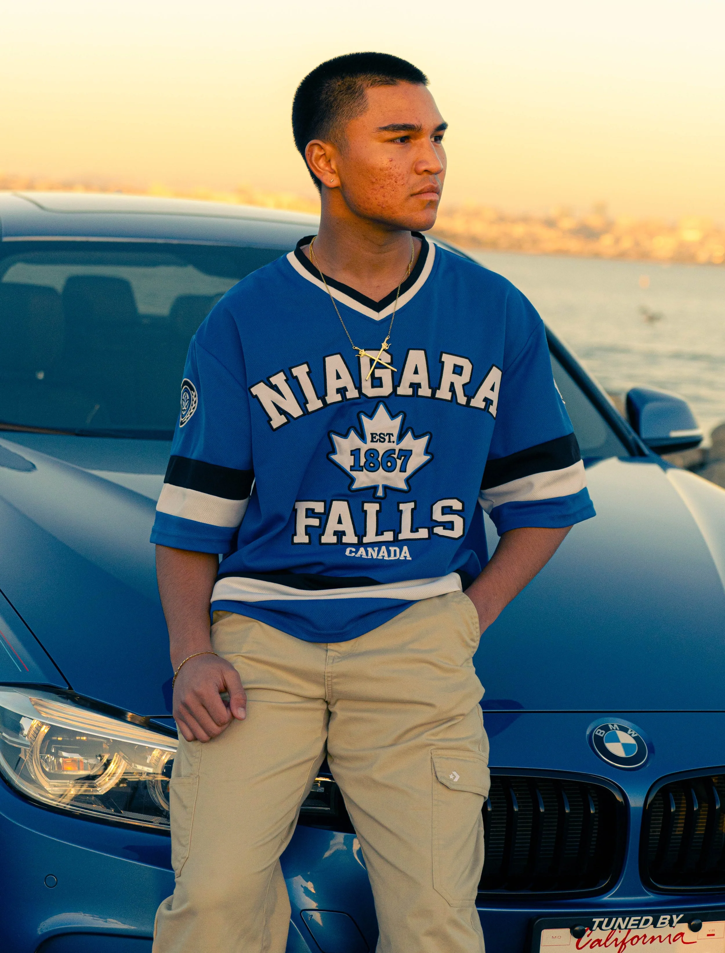 A young man stands in front of a dark blue BMW car, wearing a blue 'Niagara Falls' hockey jersey and beige cargo shorts, with a water body and blurred cityscape in the background.