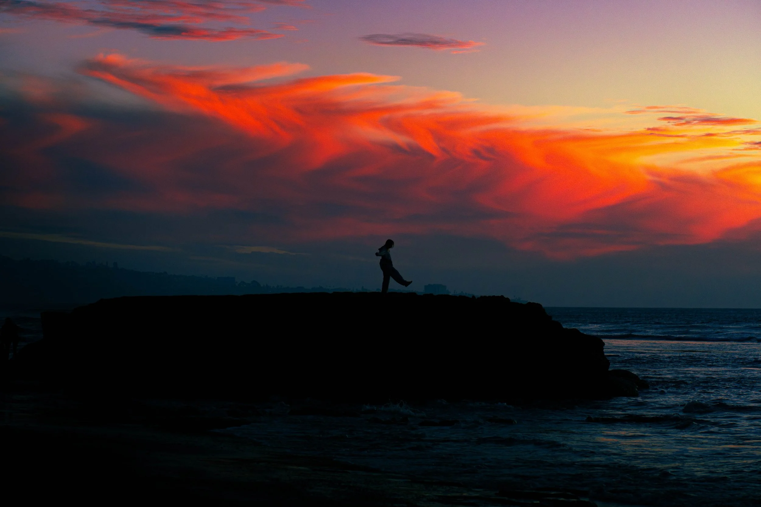 Person standing on the edge of a dark rocky shoreline at sunset with colorful orange, pink, and purple clouds in the sky.
